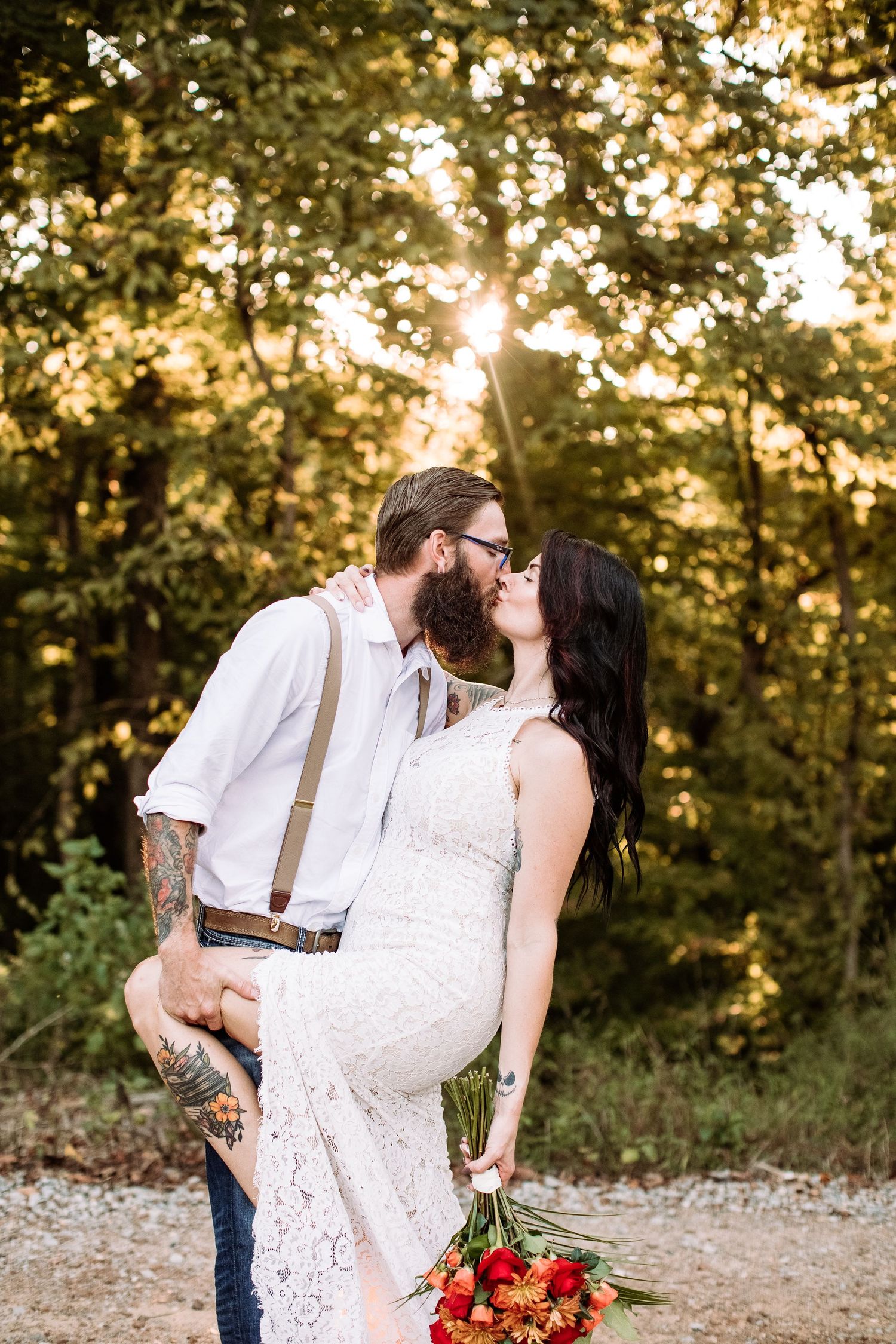 A romantic couple shares a kiss at sunset in a forest setting while holding a vibrant red bouquet.