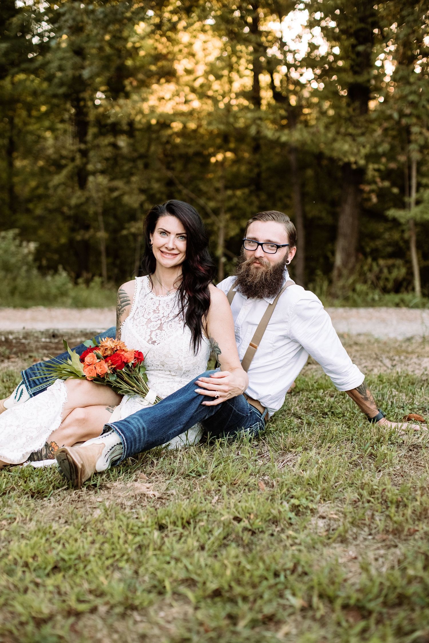 A couple in casual attire relaxes on grassy ground during an outdoor photo session at sunset.