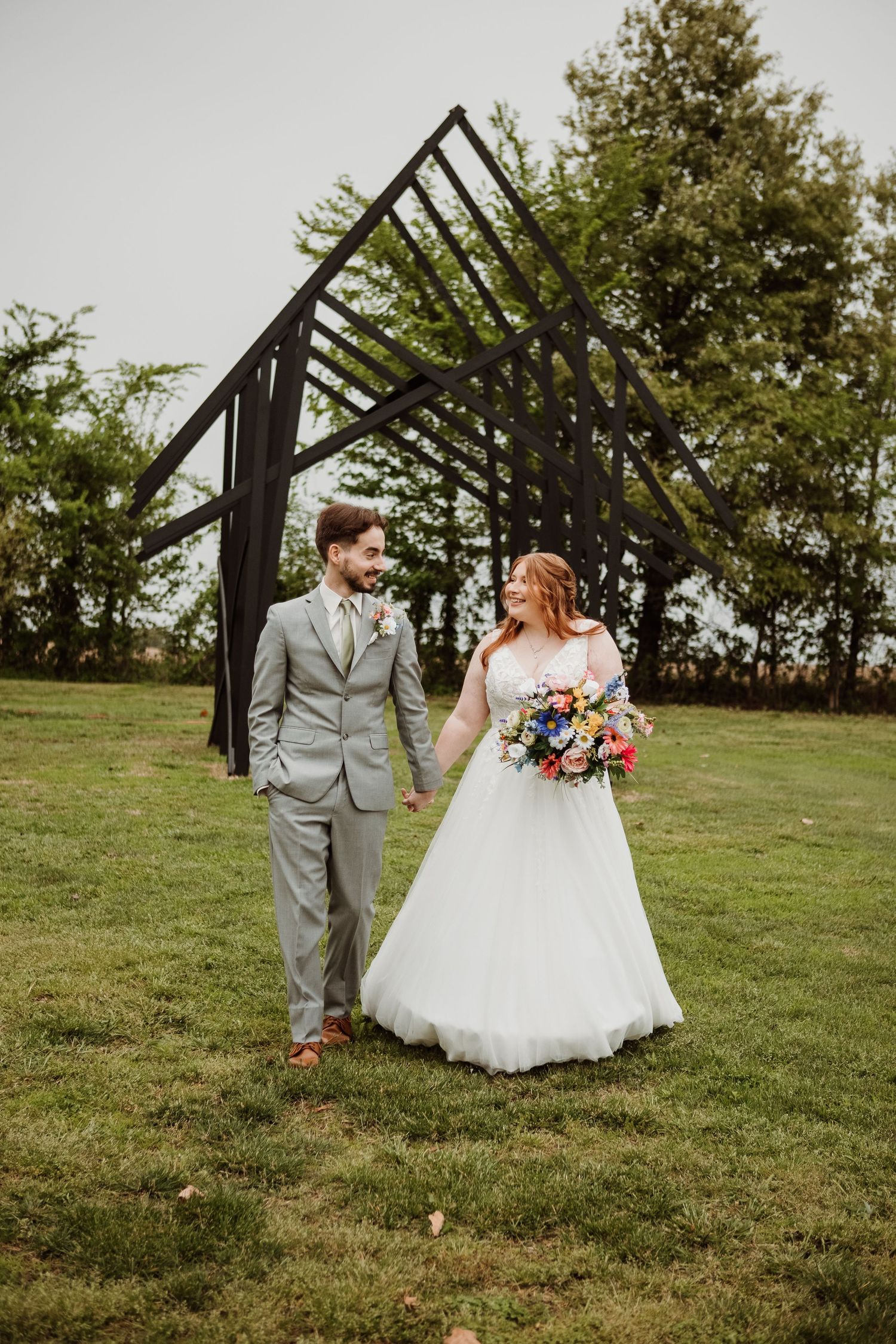 Bride and groom holding hands walking on green grass with a black metal arch backdrop on their wedding day.