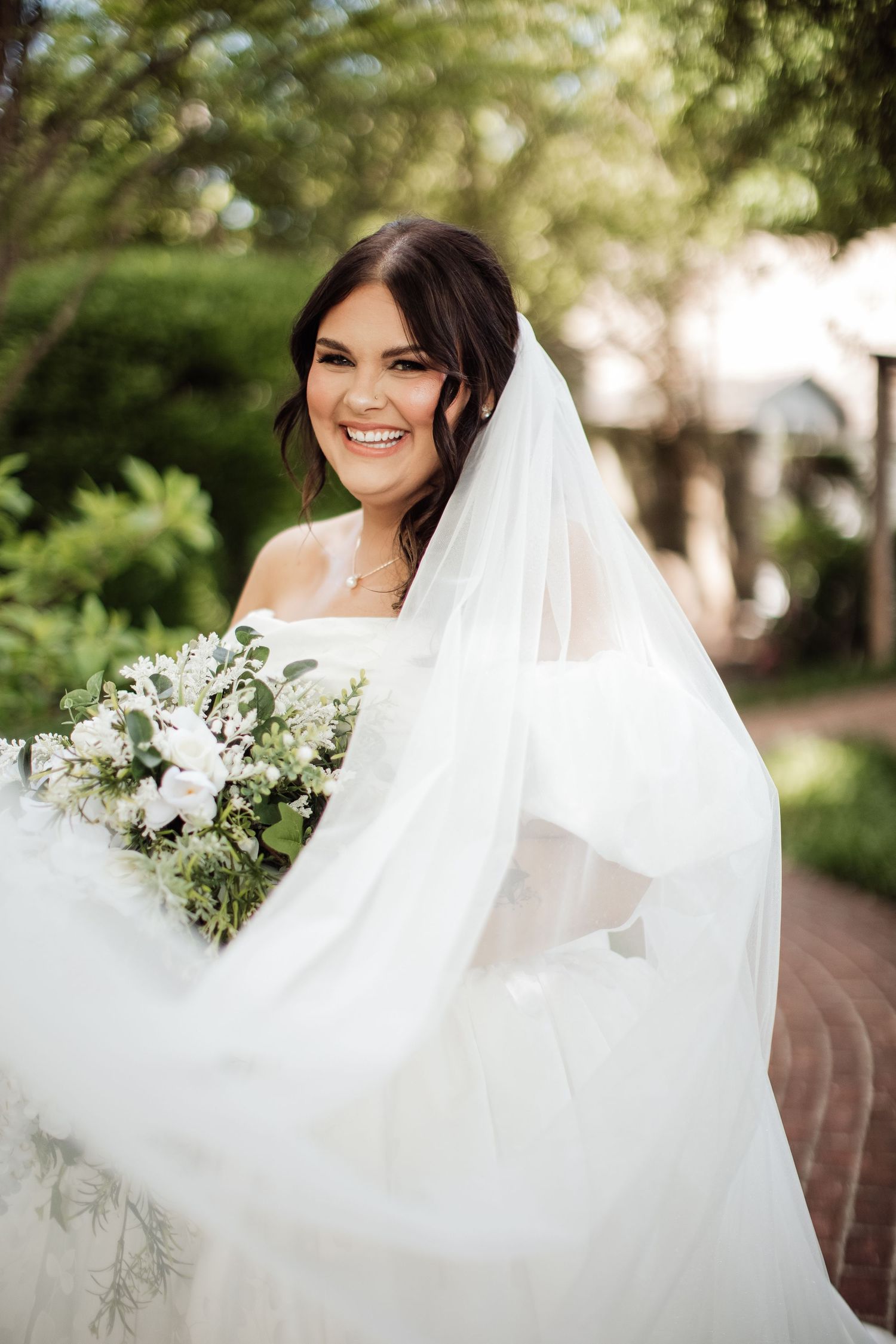 A bride in a flowing white wedding dress and veil poses outdoors with a white and green floral bouquet.