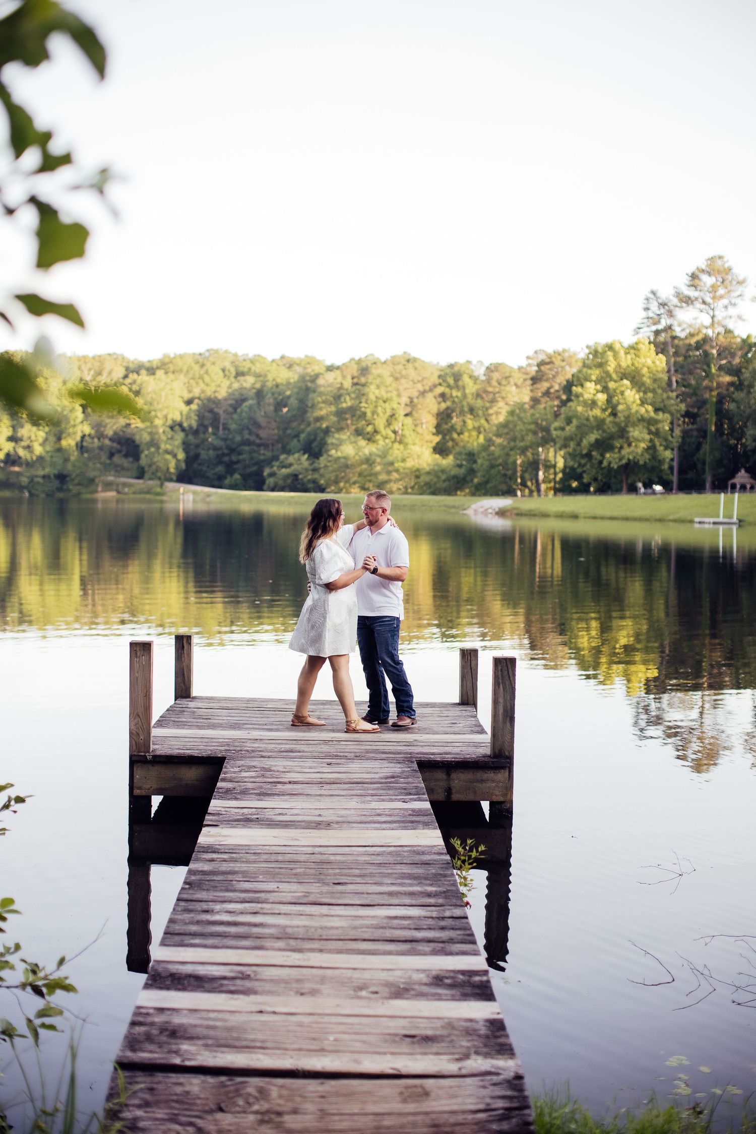 A couple shares an intimate moment on a wooden dock overlooking a serene lake surrounded by trees at sunset.
