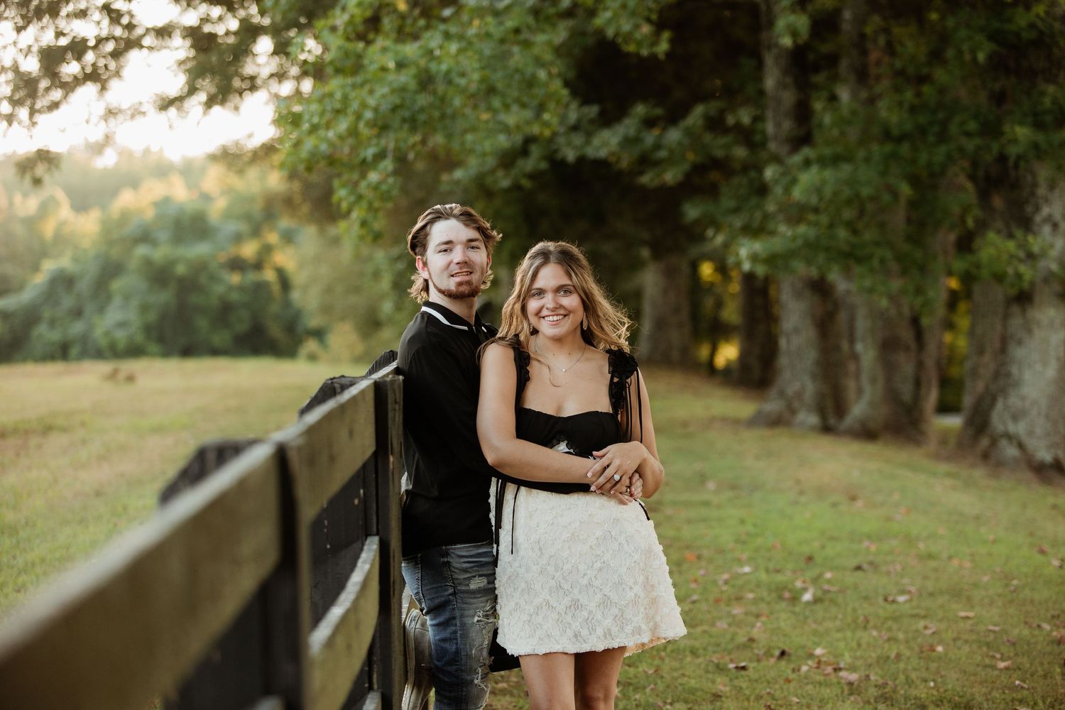 A couple poses together along a wooden fence at sunset in a scenic outdoor setting with trees in the background.