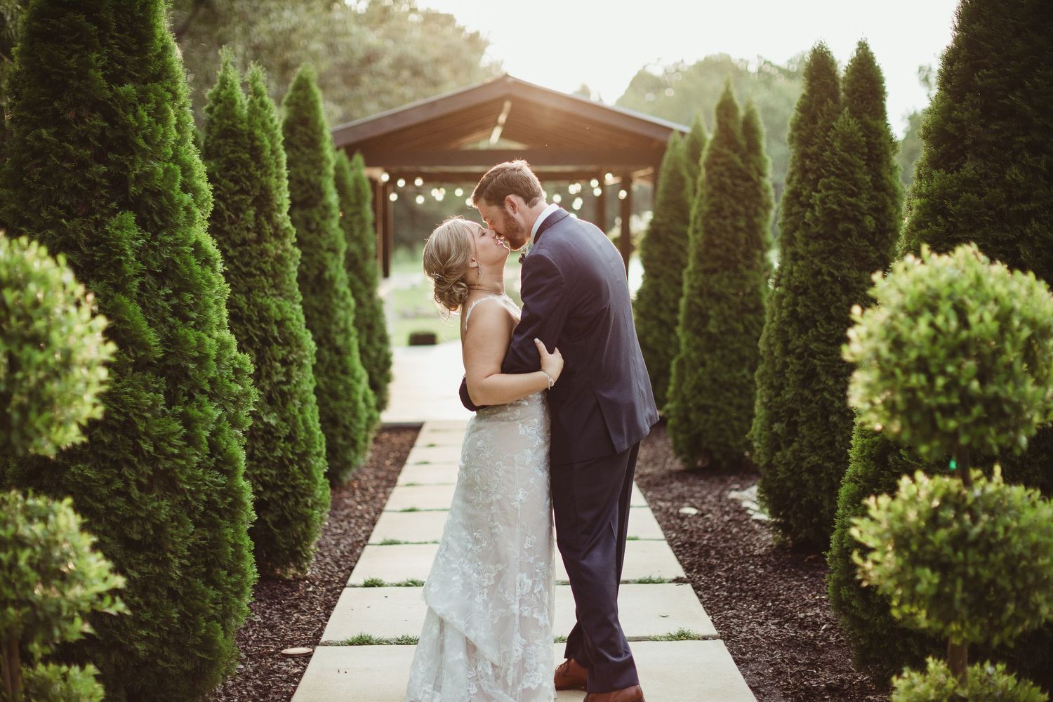A couple embraces in a romantic garden setting surrounded by tall evergreen trees and a wooden pergola.