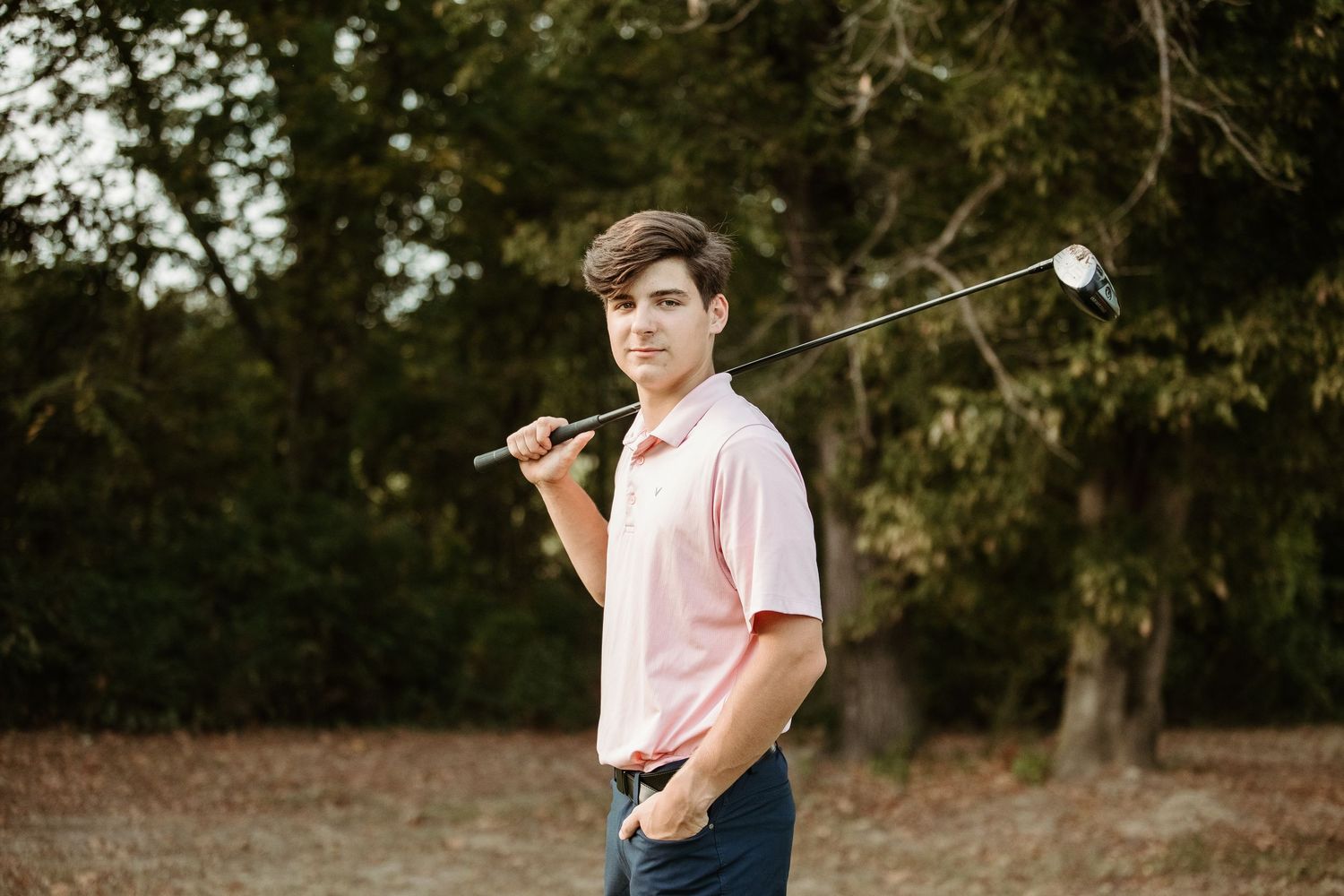 A person in a pink shirt and navy pants poses outdoors during a portrait photography session in a natural setting.