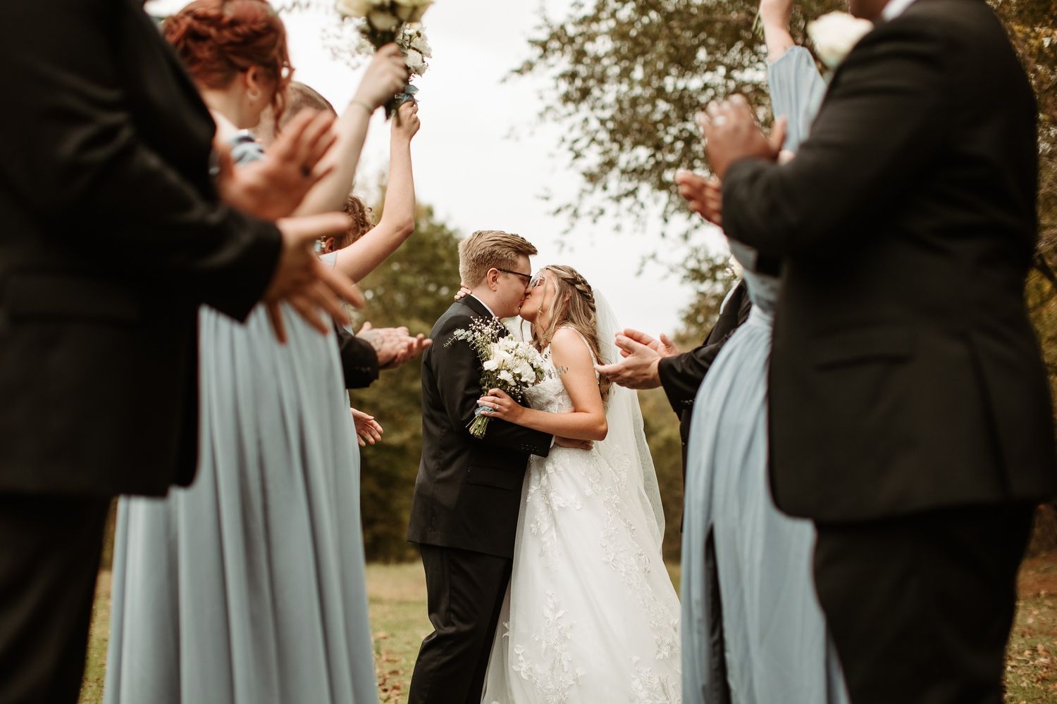 Bride and groom share a kiss surrounded by bridesmaids in blue dresses and groomsmen during outdoor wedding ceremony.