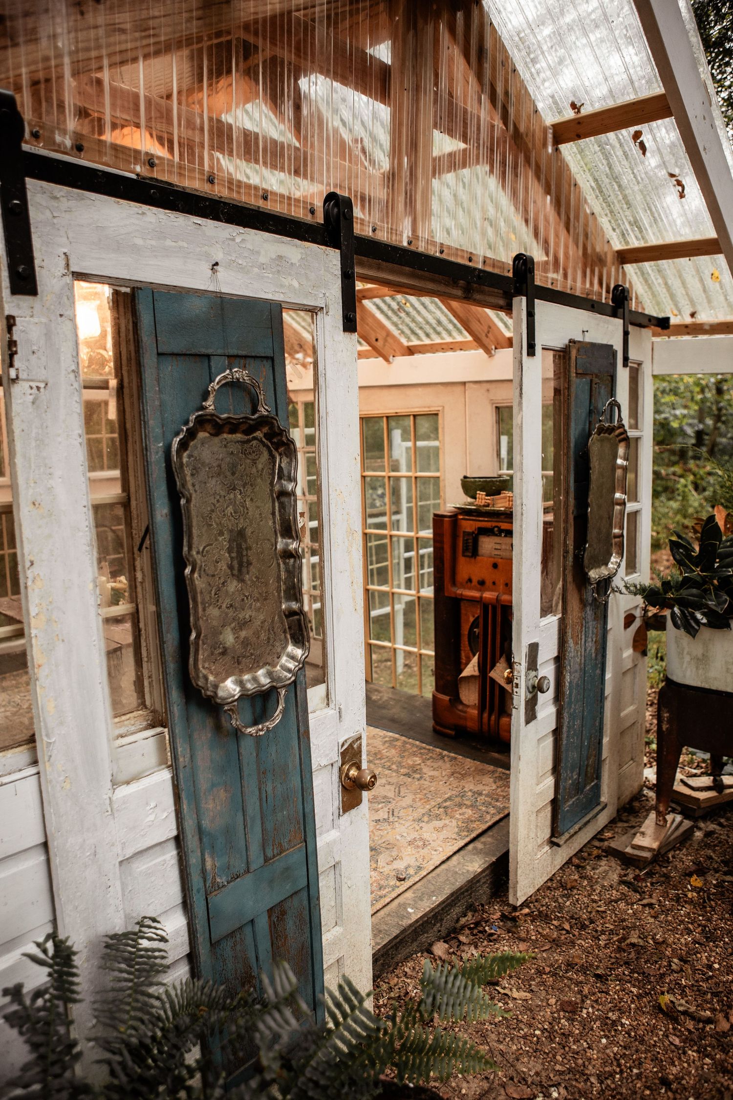 Rustic greenhouse built from salvaged white and teal windows and doors, with ornate silver trays hung as decor and ferns growing outside.