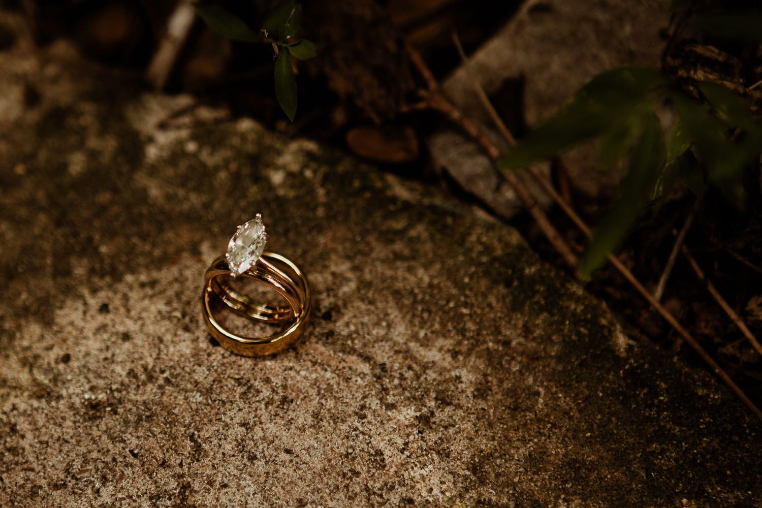 Gold wedding bands and diamond engagement ring stacked on rustic stone surface with natural greenery background.
