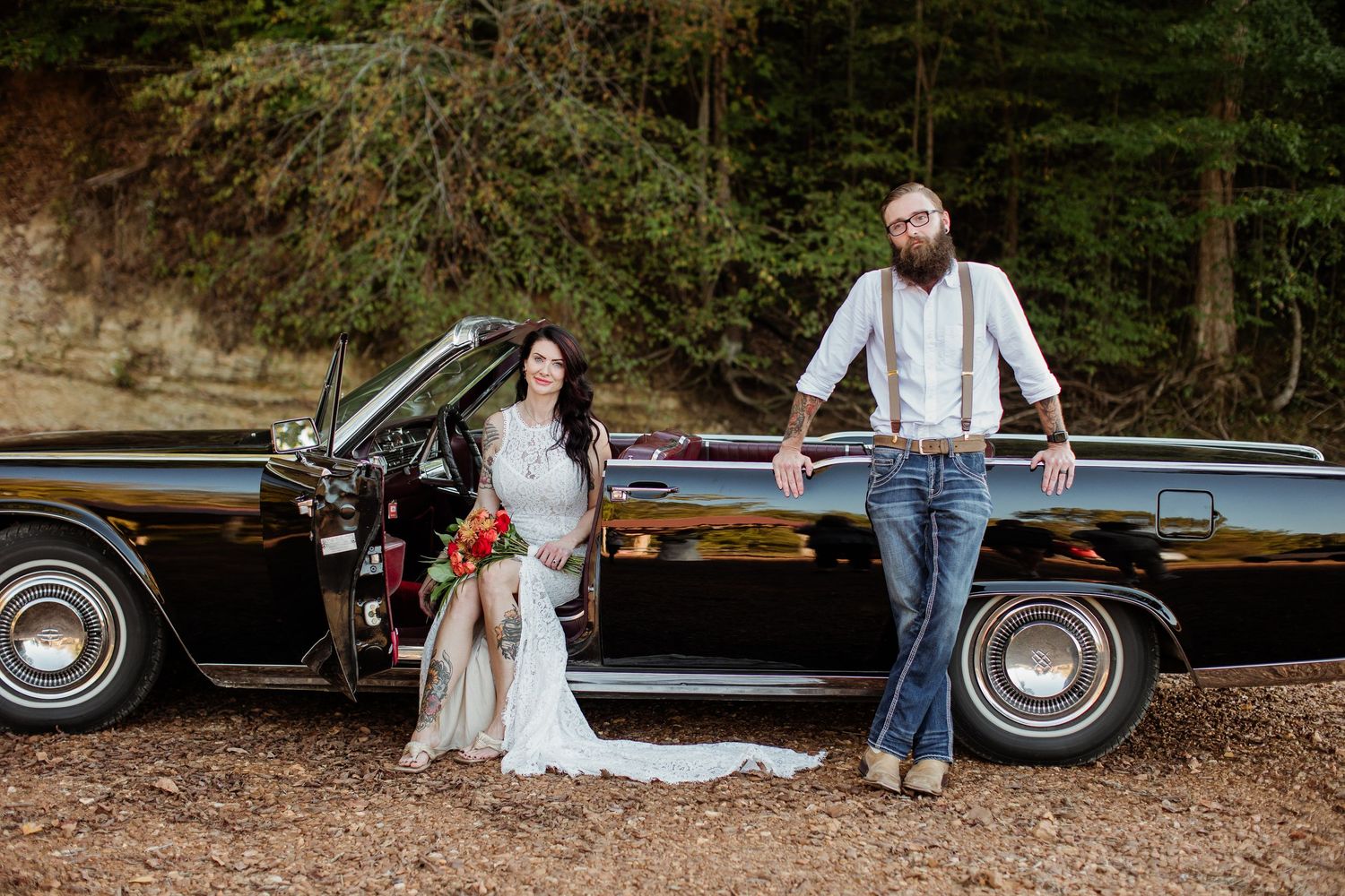 A couple sits on a blanket near a classic black car in a wooded outdoor setting.