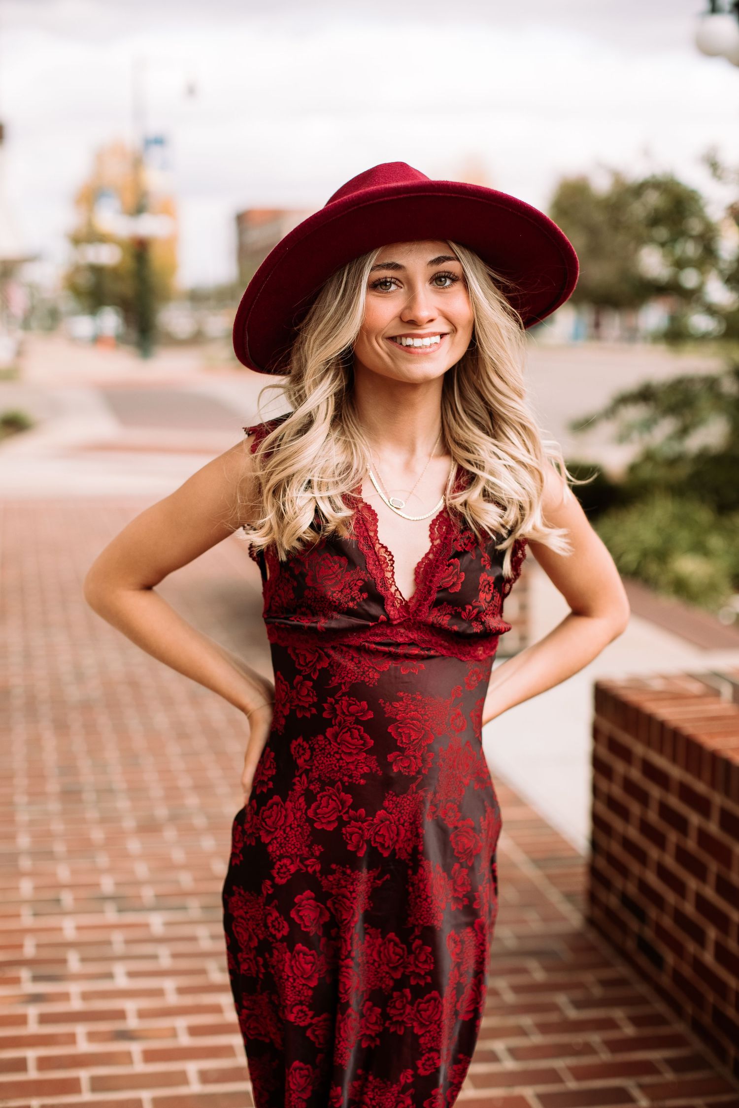 Smiling blonde woman in a red floral dress and matching wide-brim hat posing outdoors on a brick walkway.