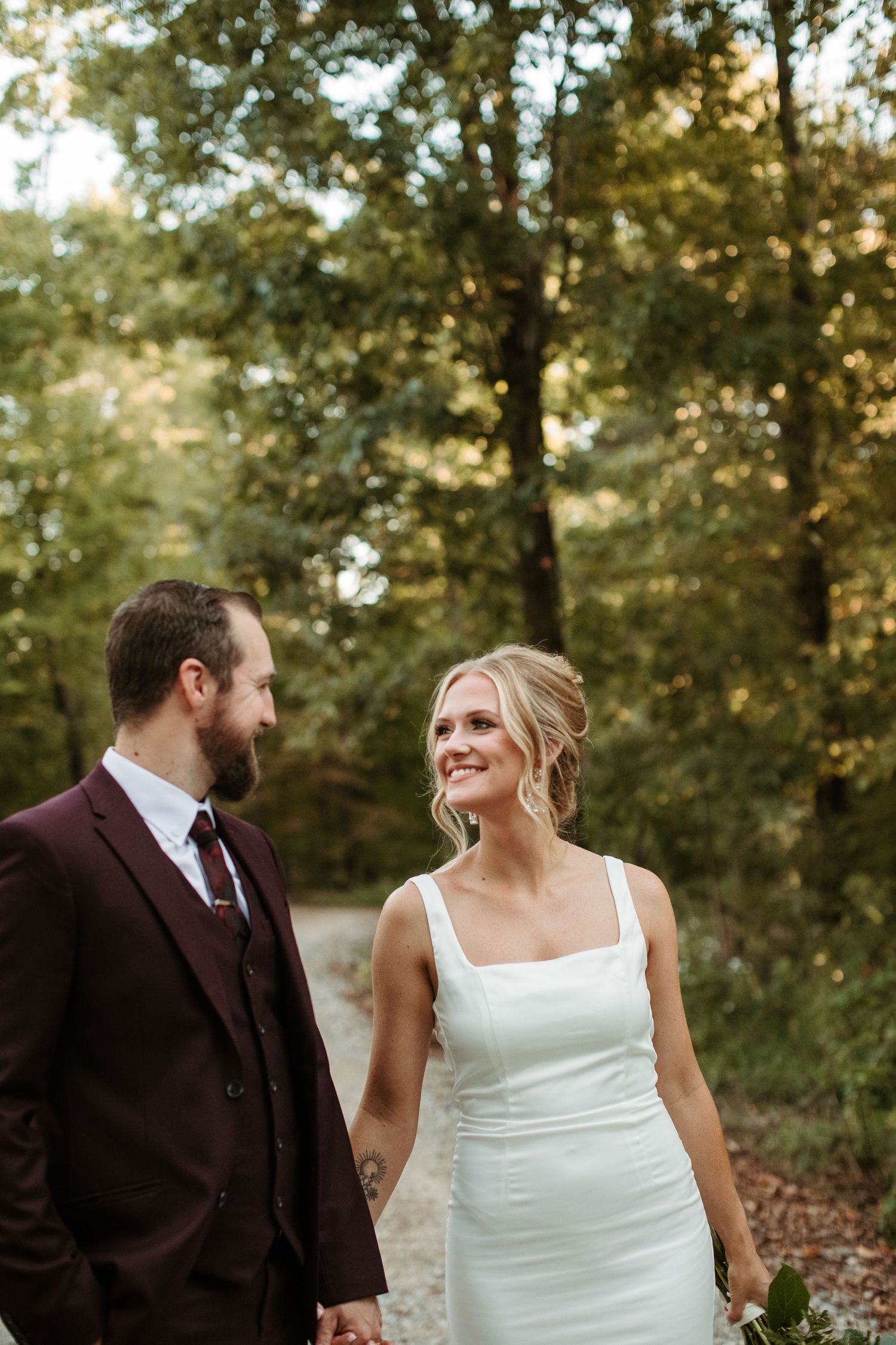A couple in wedding attire share joyful laughter while standing together in a sunlit wooded area.