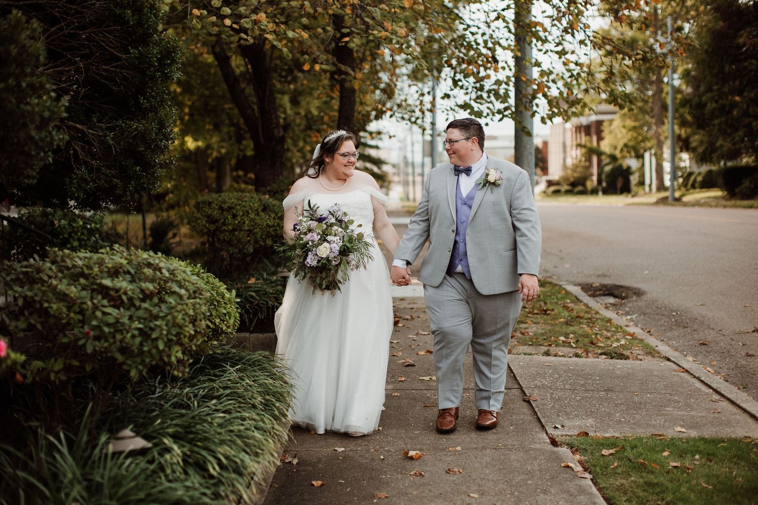 A happy couple shares a joyful moment together on their wedding day, standing on a tree-lined sidewalk.