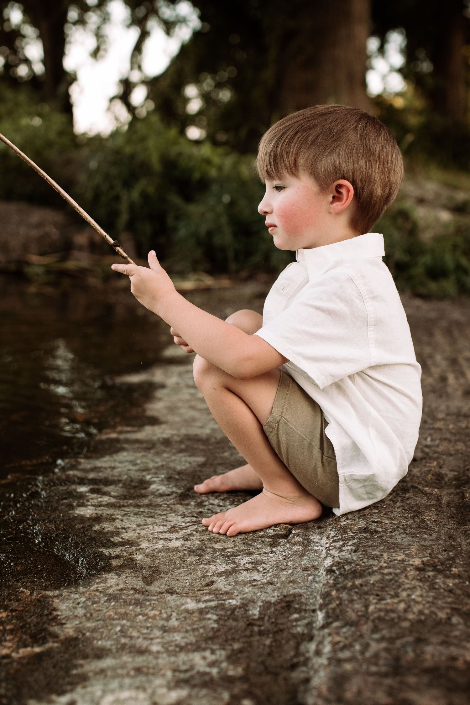 A young child in white shirt and khaki shorts sits barefoot on a rock while fishing in a stream during golden hour.