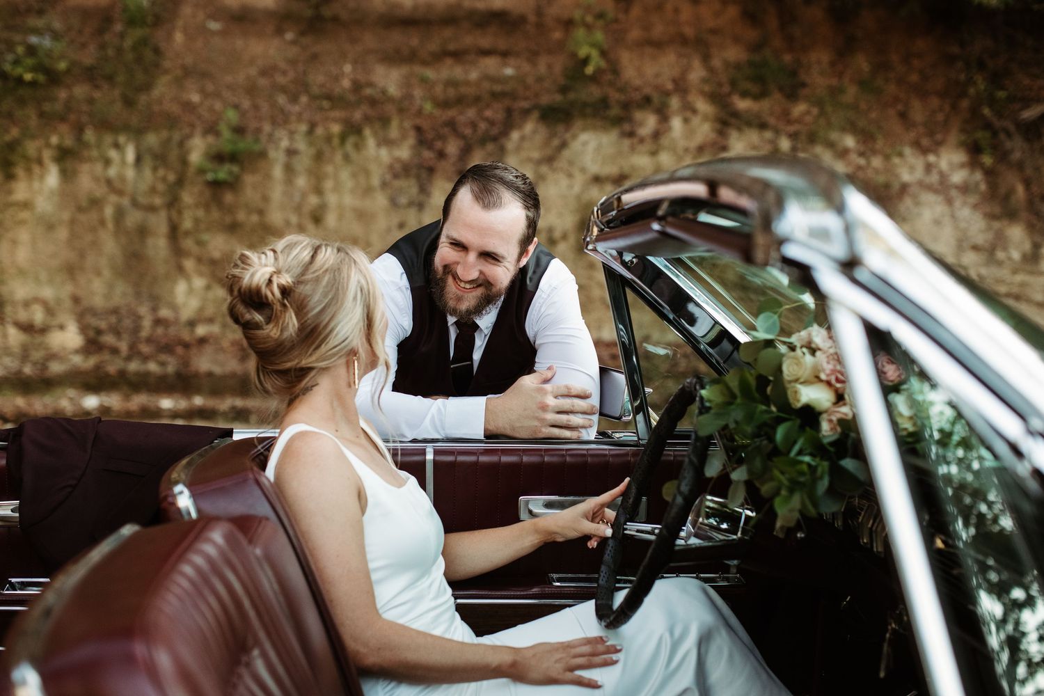 Wedding party members use mobile phones while sitting in a vintage car decorated with greenery.