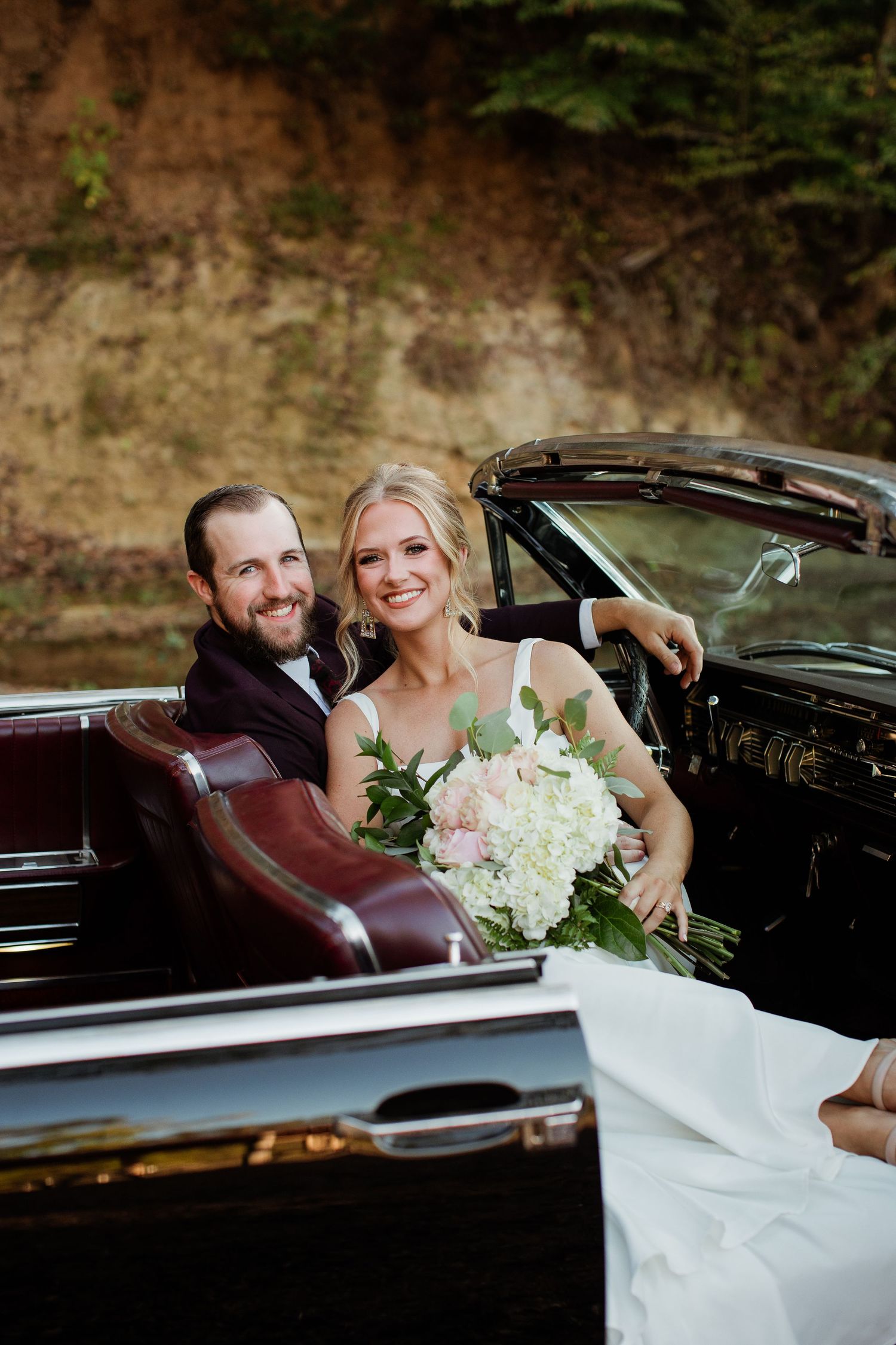 A couple sits together in a vintage car with burgundy leather interior during a wedding celebration.
