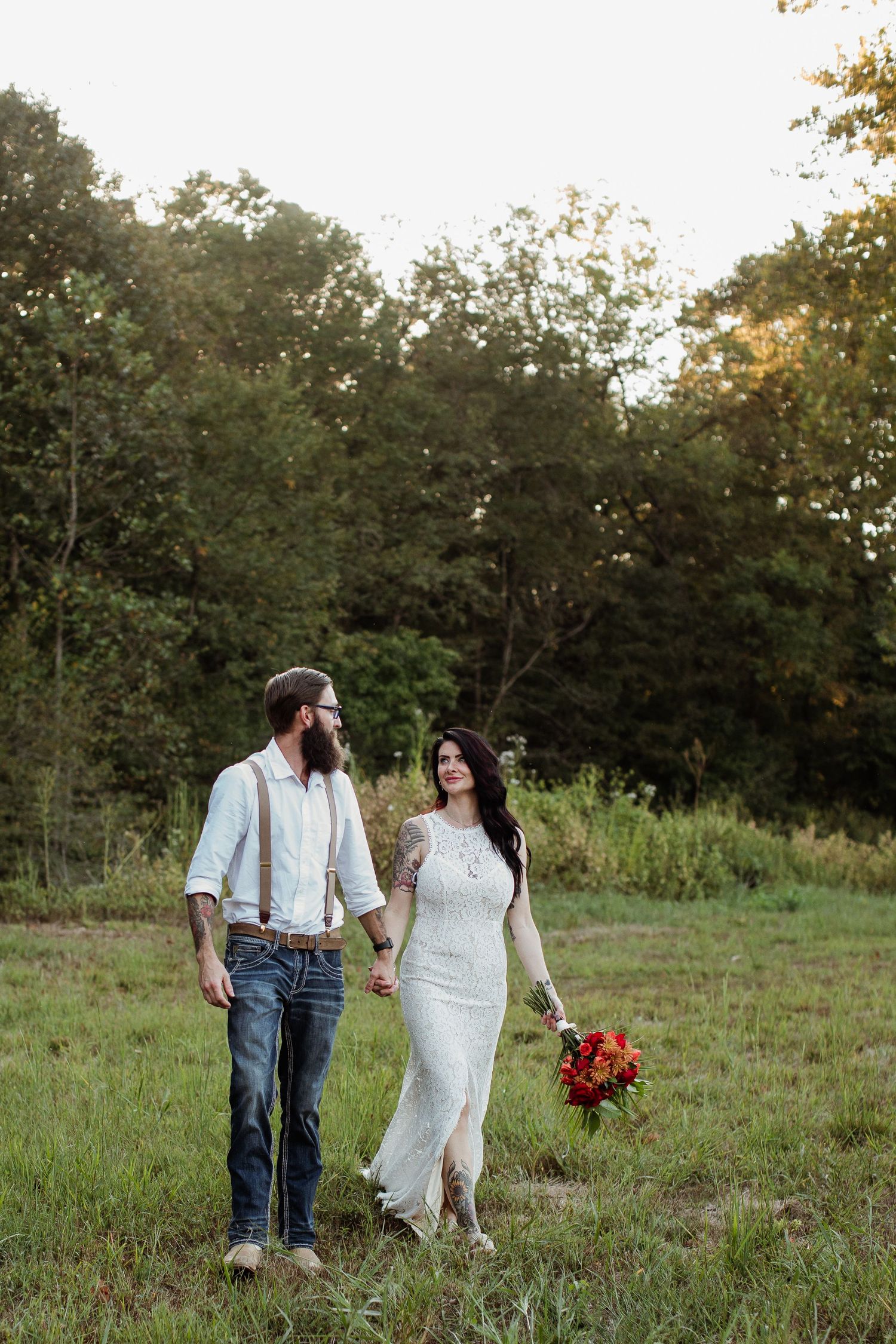 A couple walks hand in hand through a sunlit meadow with trees in the background during golden hour.