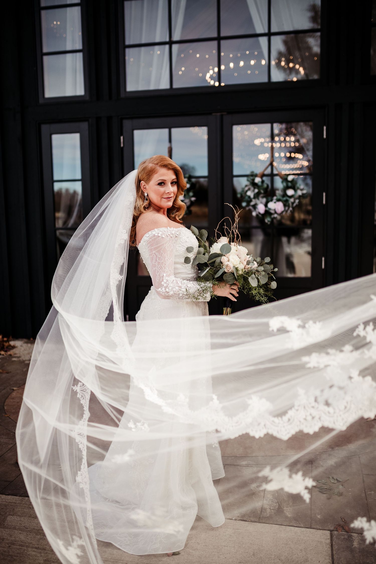 A bride in a white lace gown stands elegantly as her long veil flows dramatically in the wind against dark windows.
