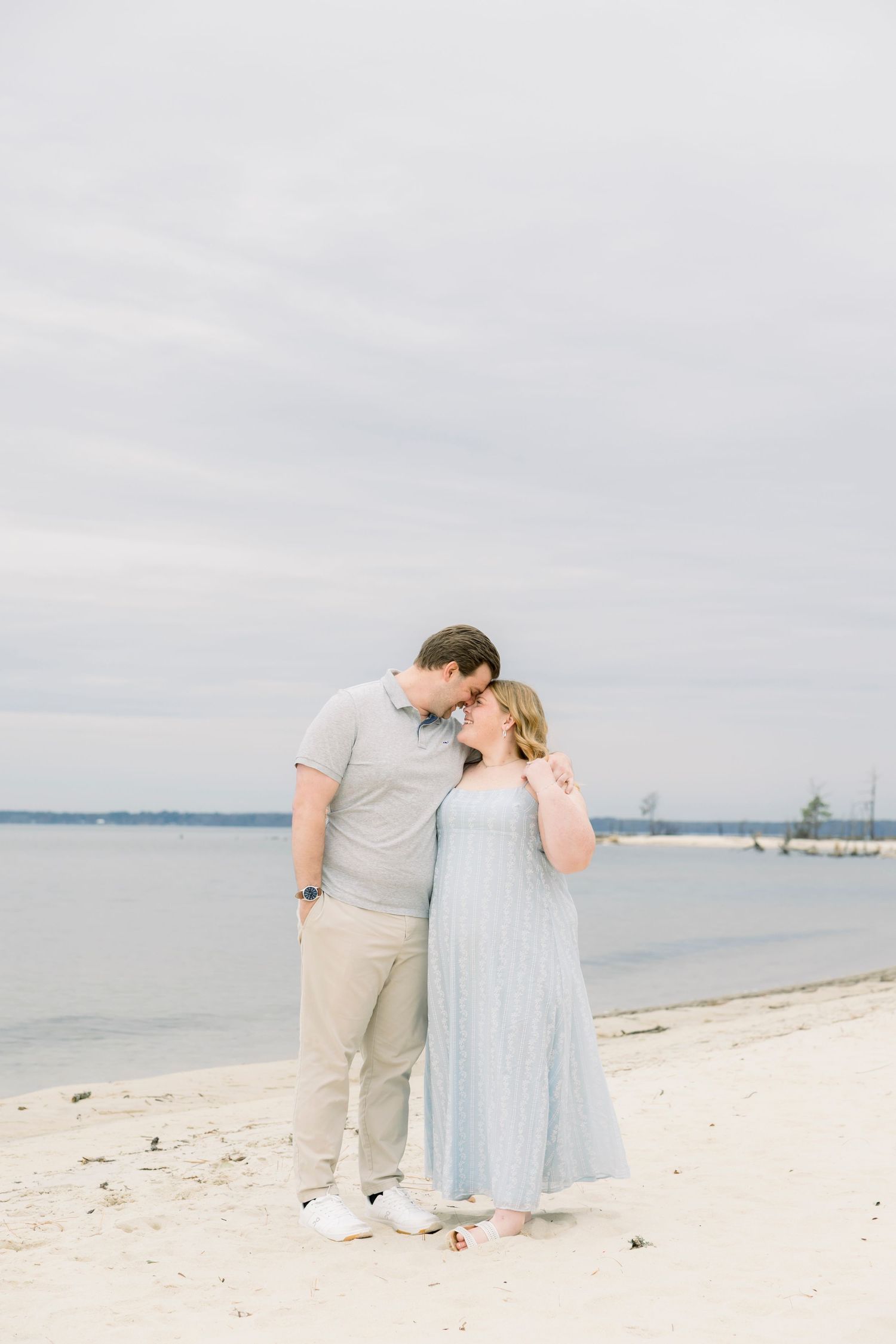 A couple shares an intimate moment while standing on a peaceful beach with water and cloudy skies in the background.