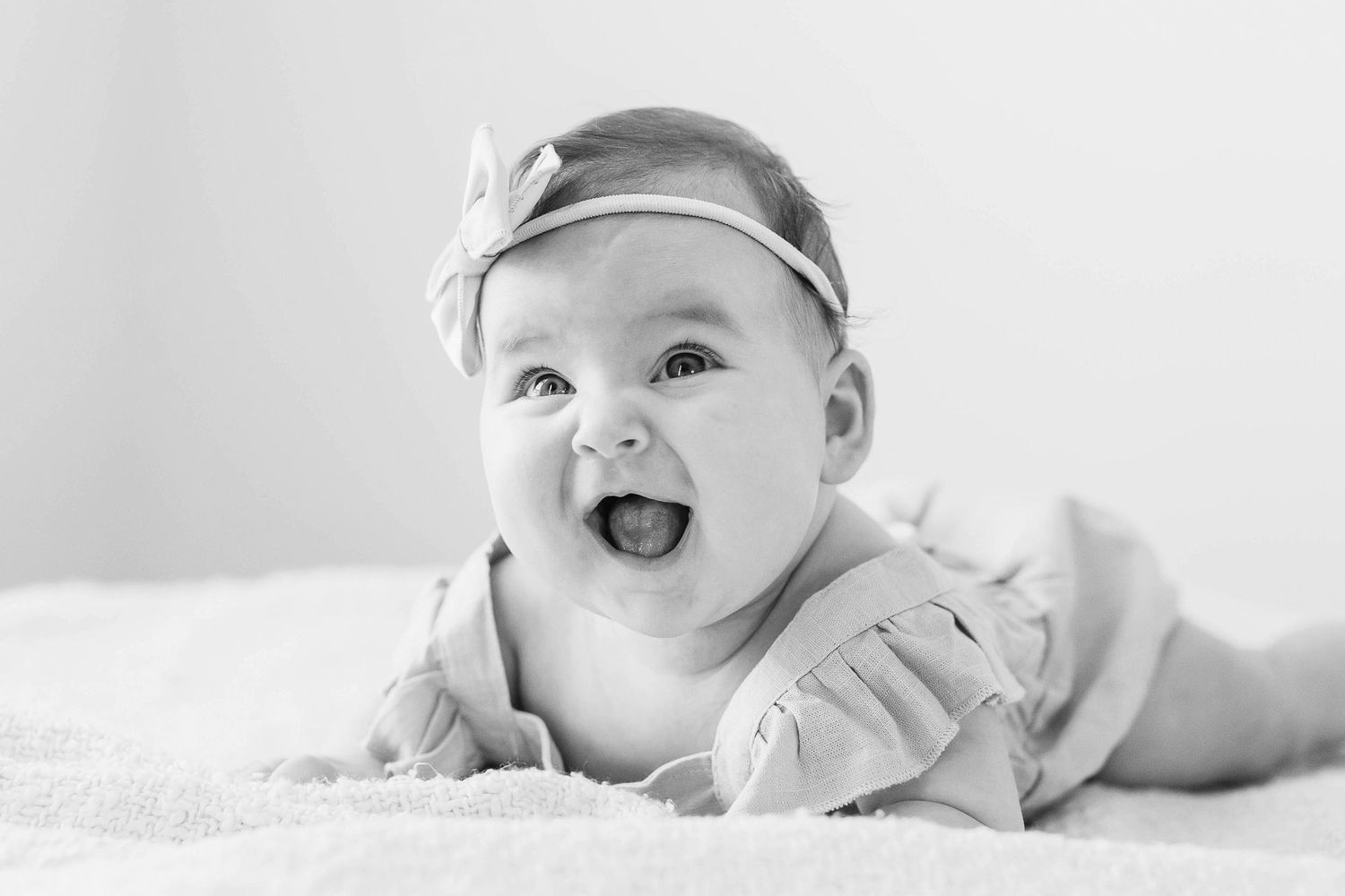 Black and white photo of a happy baby with a bow headband lying on their stomach.