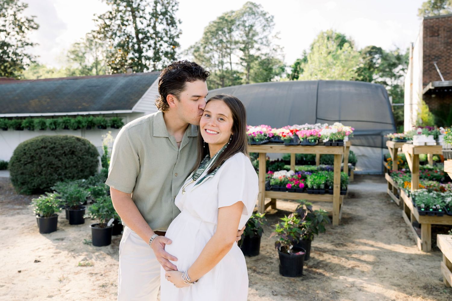 A couple shares a tender maternity moment surrounded by colorful flowers and plants at a garden nursery.