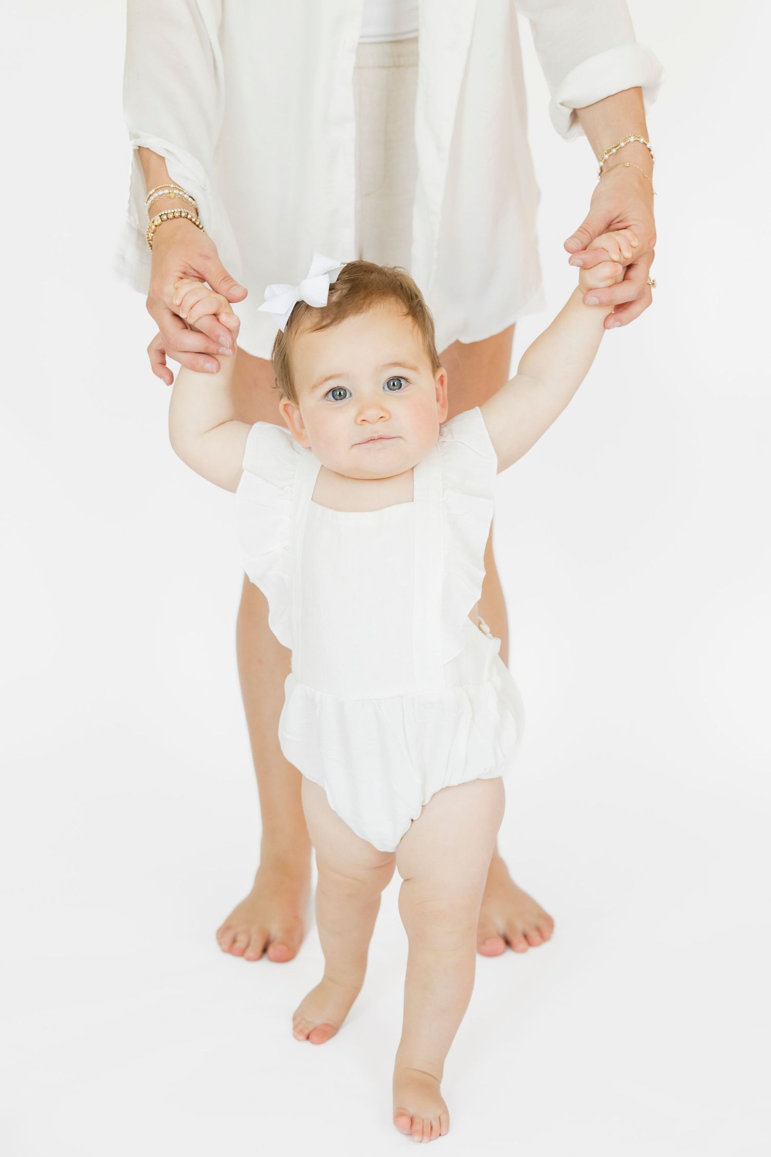 Baby in white outfit standing with support against white background.