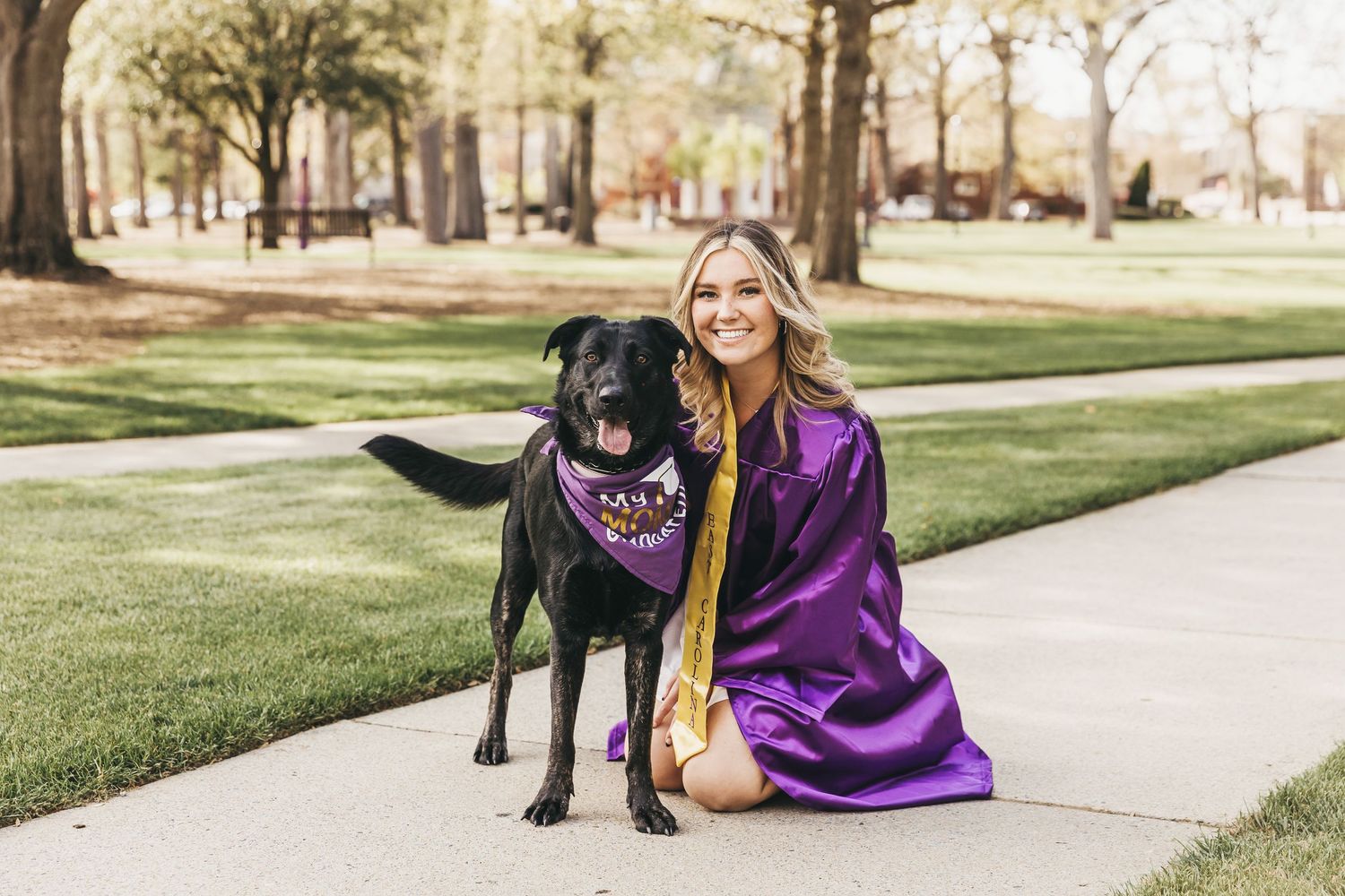 Person in purple graduation gown kneeling next to black dog wearing bandana outdoors.