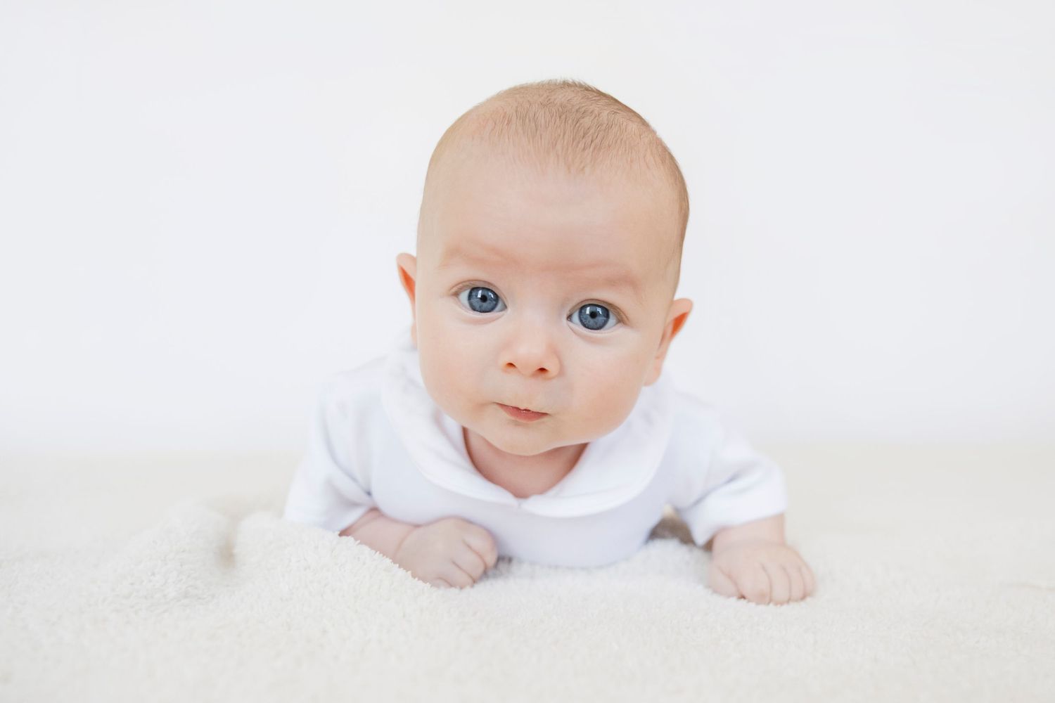 Adorable blue-eyed baby in white onesie doing tummy time on a soft white blanket.