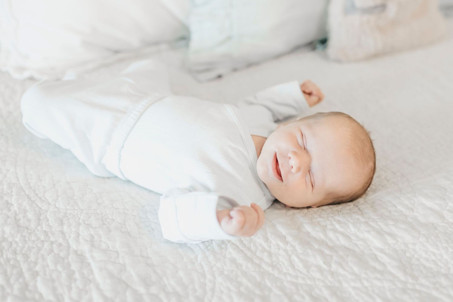 A newborn baby wearing white sleeps peacefully on soft white bedding.