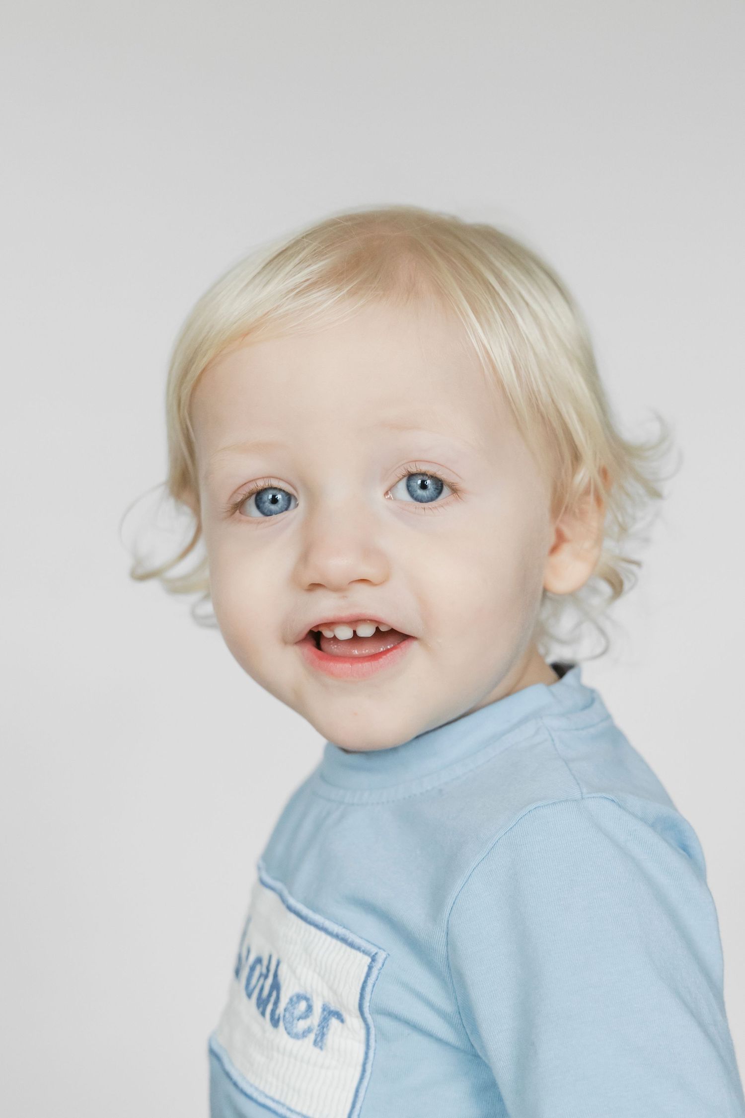 A toddler with bright blue eyes wearing a light blue shirt smiles at the camera.