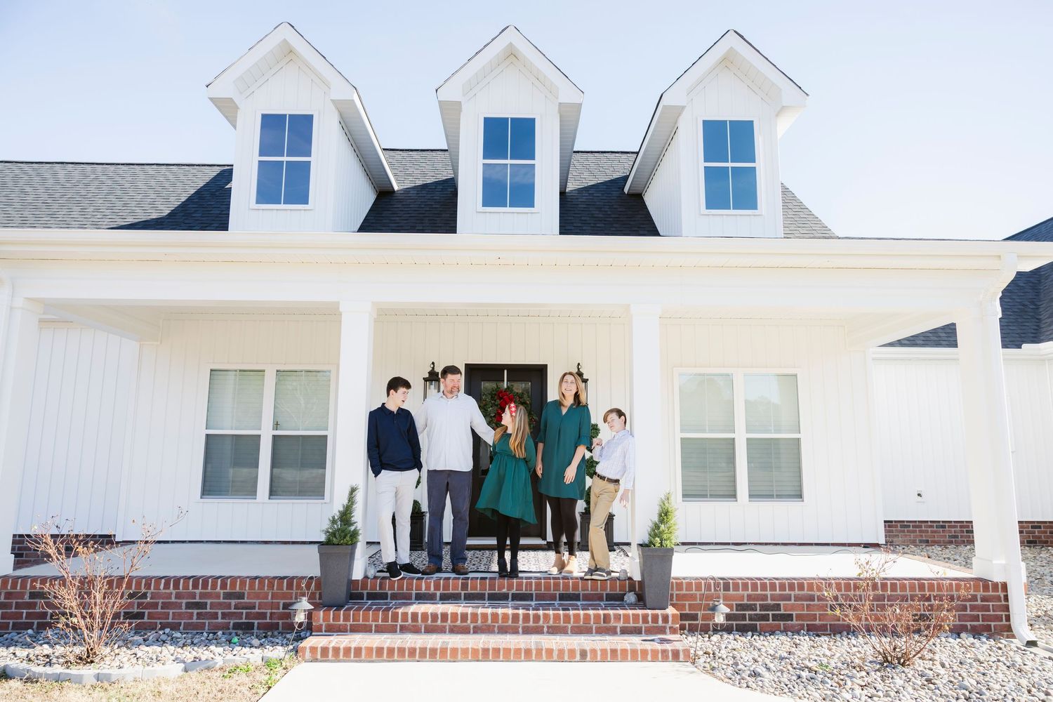 Family of five posing on front porch of modern white farmhouse with brick steps and dormer windows.