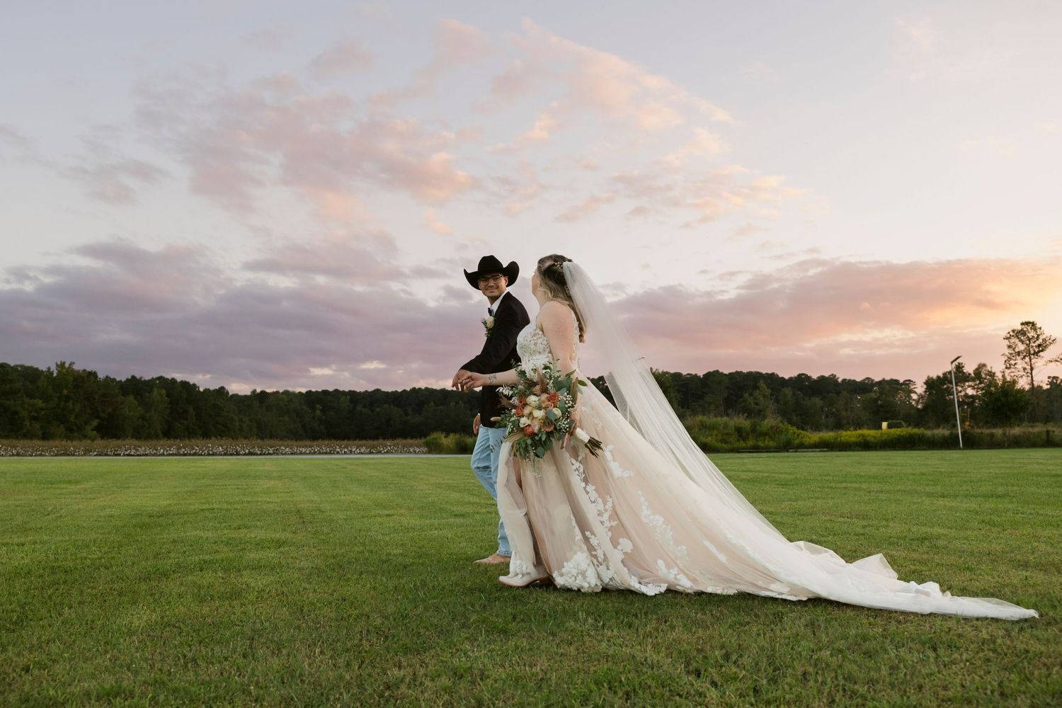 A bride in a flowing white gown stands in a golden field at sunset.