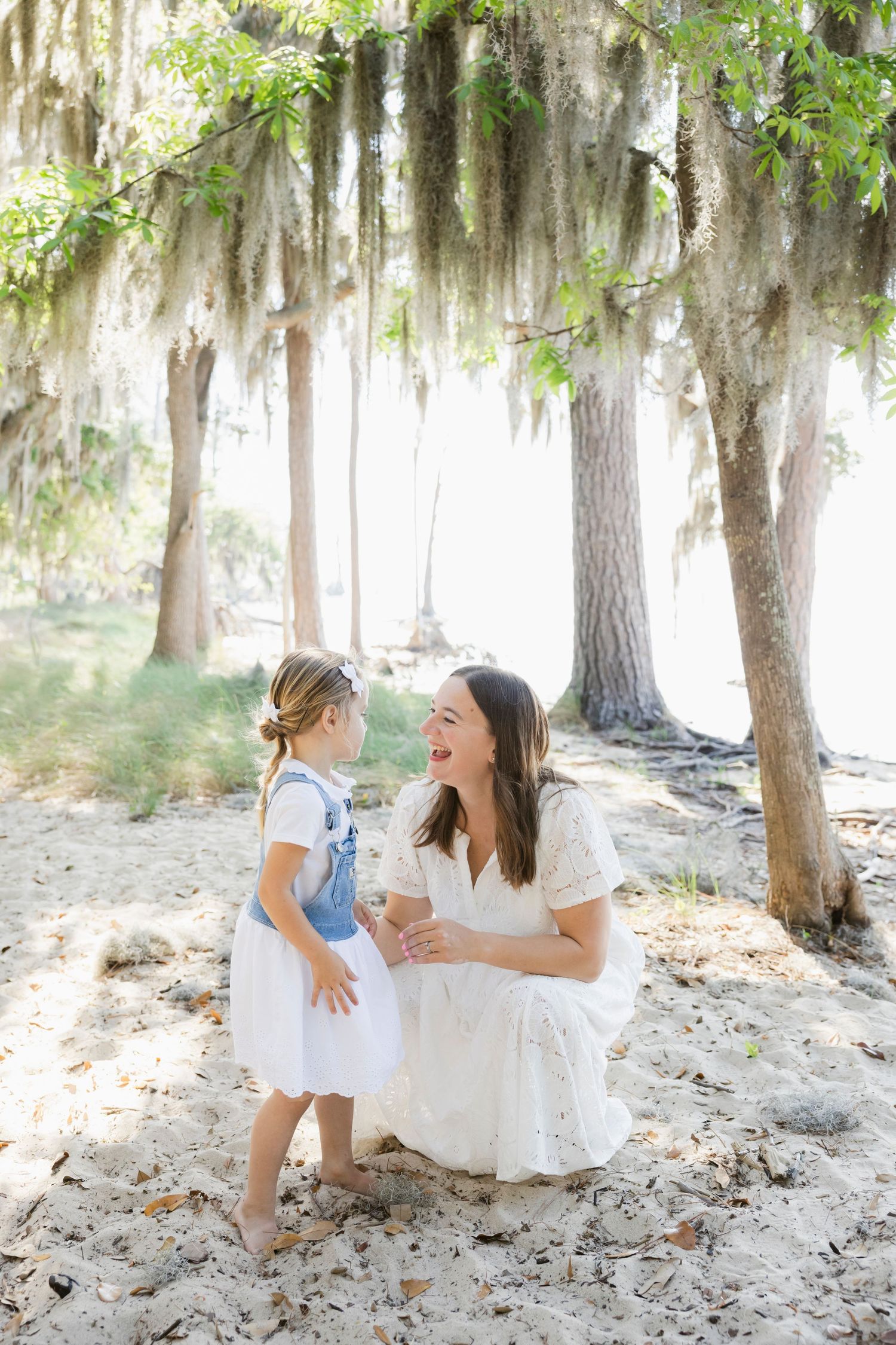 A mother crouches to smile at her young daughter under moss-draped trees on a sandy path, both wearing white outfits.