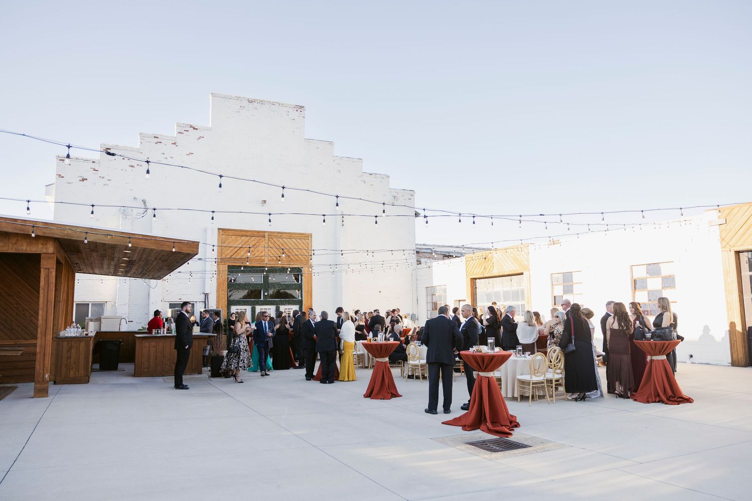 A panoramic view of an outdoor event space with white walls and string lights shows guests gathered around cocktail tables.