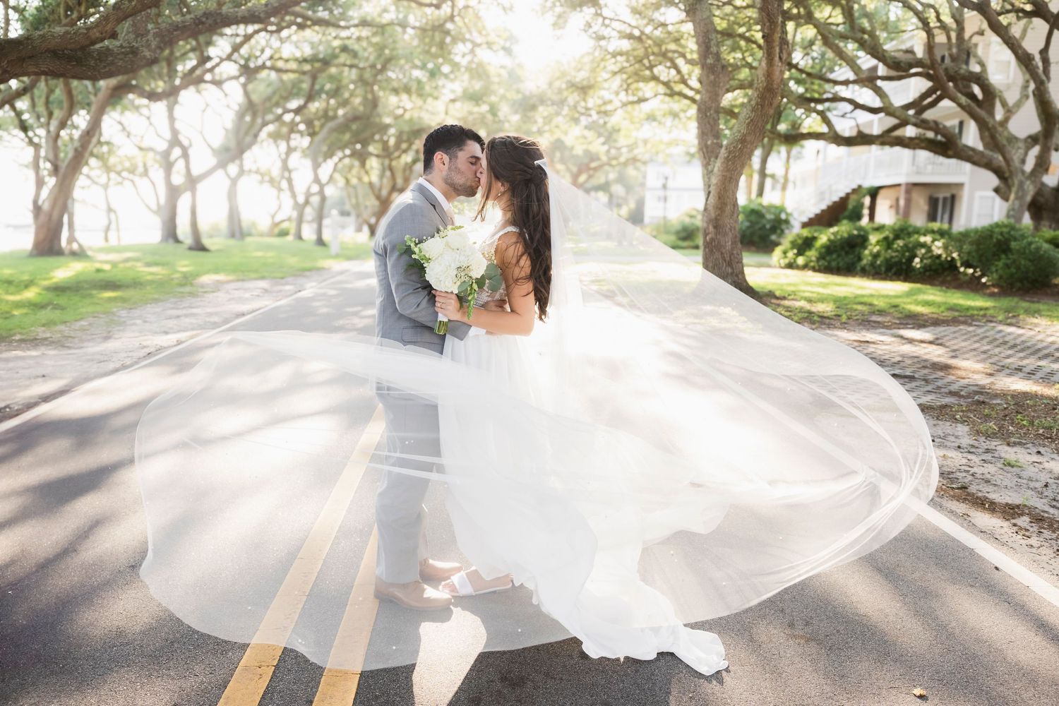Bride and groom share a romantic kiss on a sunlit road, her flowing white veil billowing gracefully in the breeze.