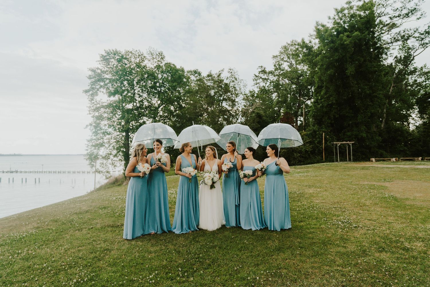 Wedding party in light blue dresses and dark suits posing on grassy hillside.
