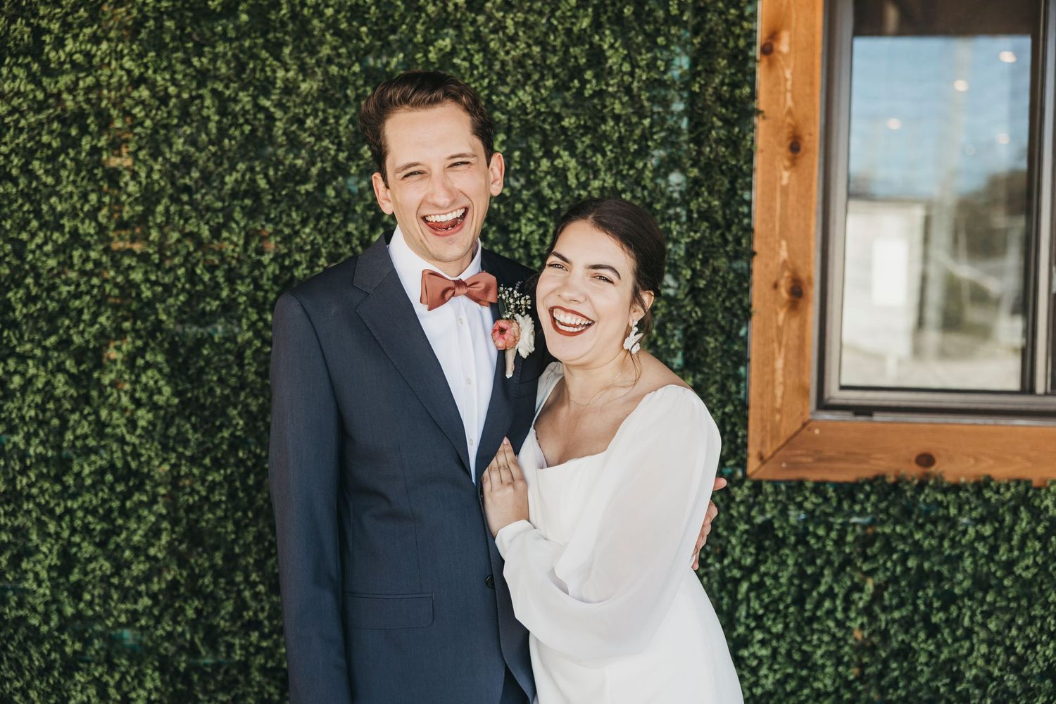 Wedding party in white and rust-colored dresses poses against a green hedge wall with wooden beams.
