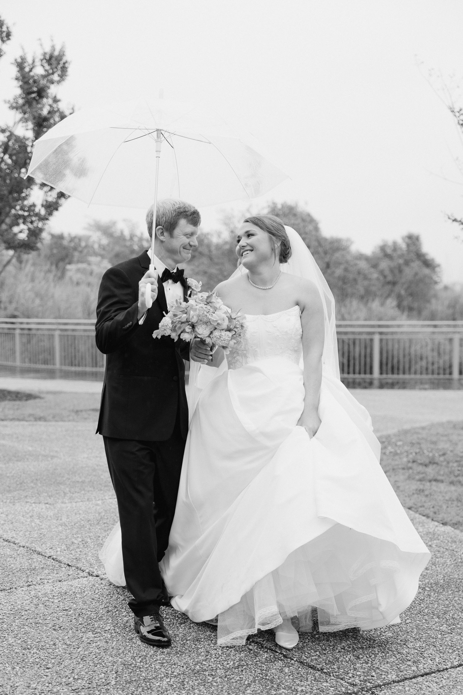 Black and white photo of smiling bride and groom walking under clear umbrella on rainy wedding day.