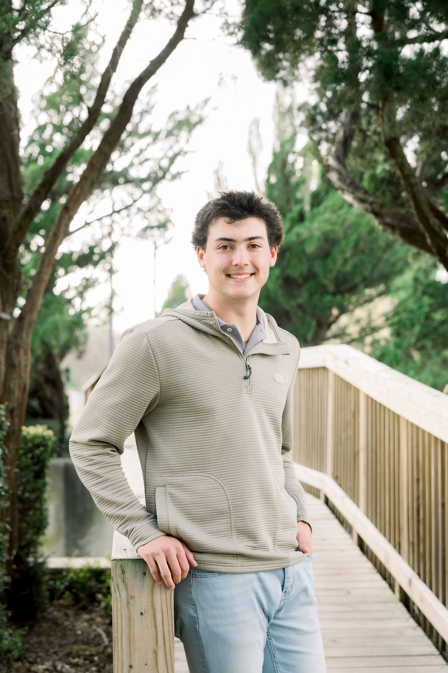Young man in a gray hoodie smiling on a wooden bridge surrounded by lush green trees.