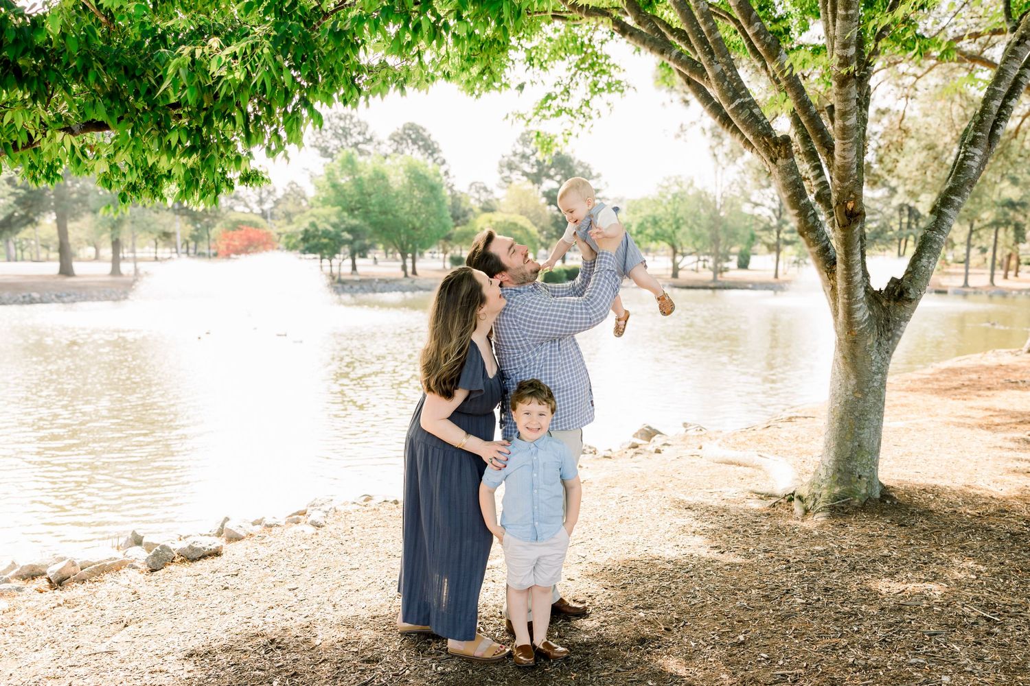 Family of four enjoying a sunny day by a lake, with dad lifting baby girl while mom and son smile nearby.