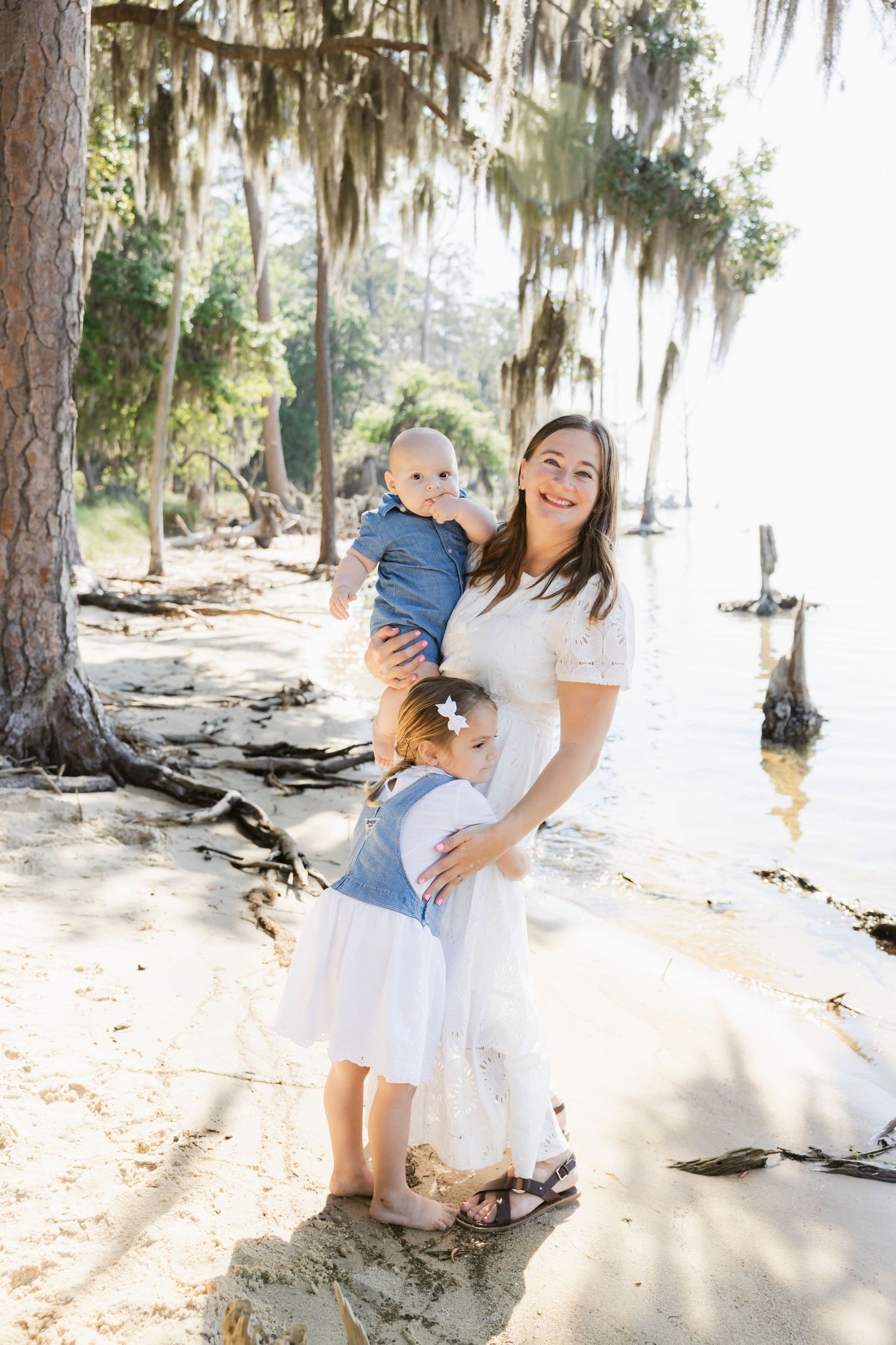 A smiling mother in a white dress holds a baby while her young daughter hugs her near a sunlit lakeside.