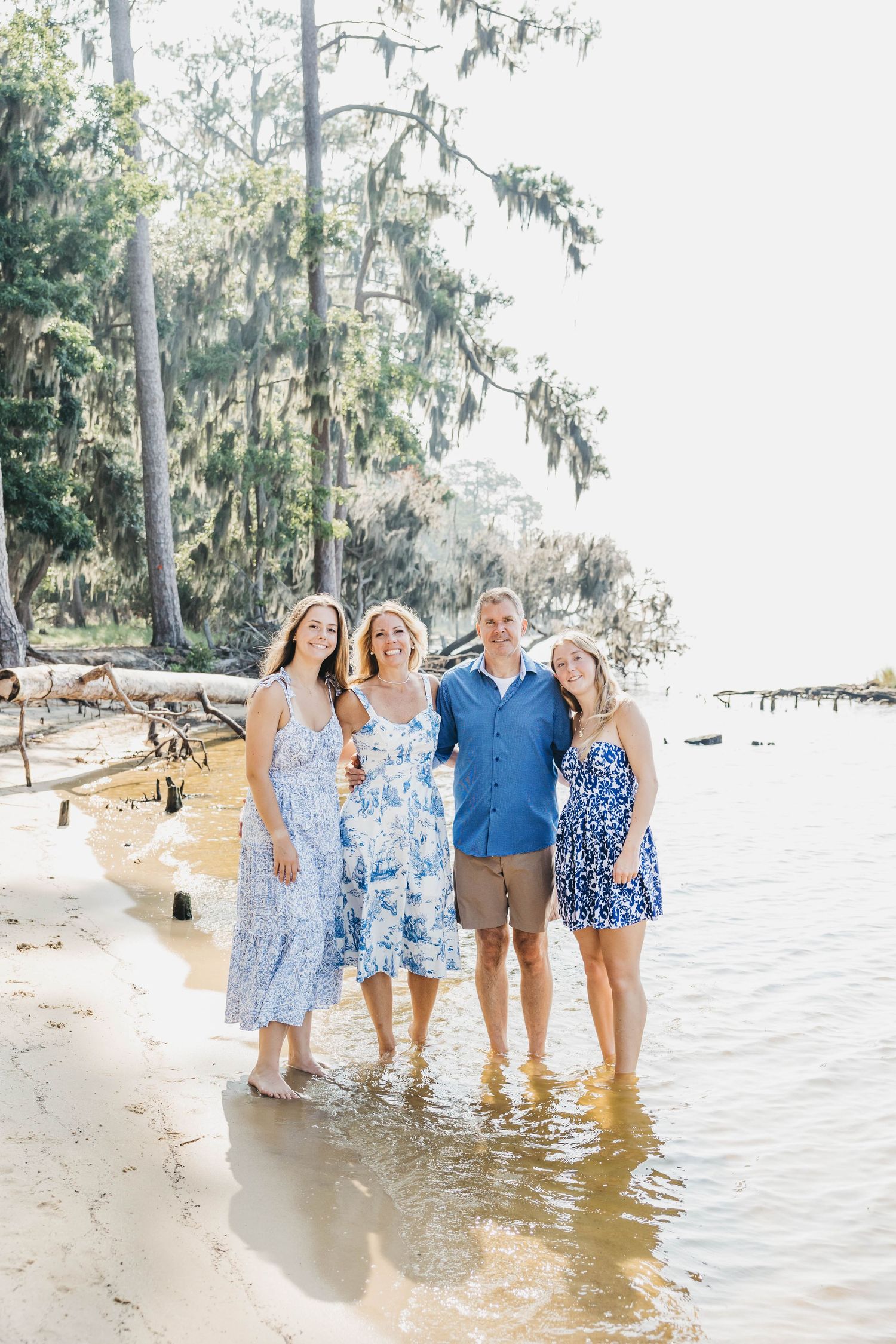 Family group wearing coordinated blue outfits poses on a sunny beach with Spanish moss trees.