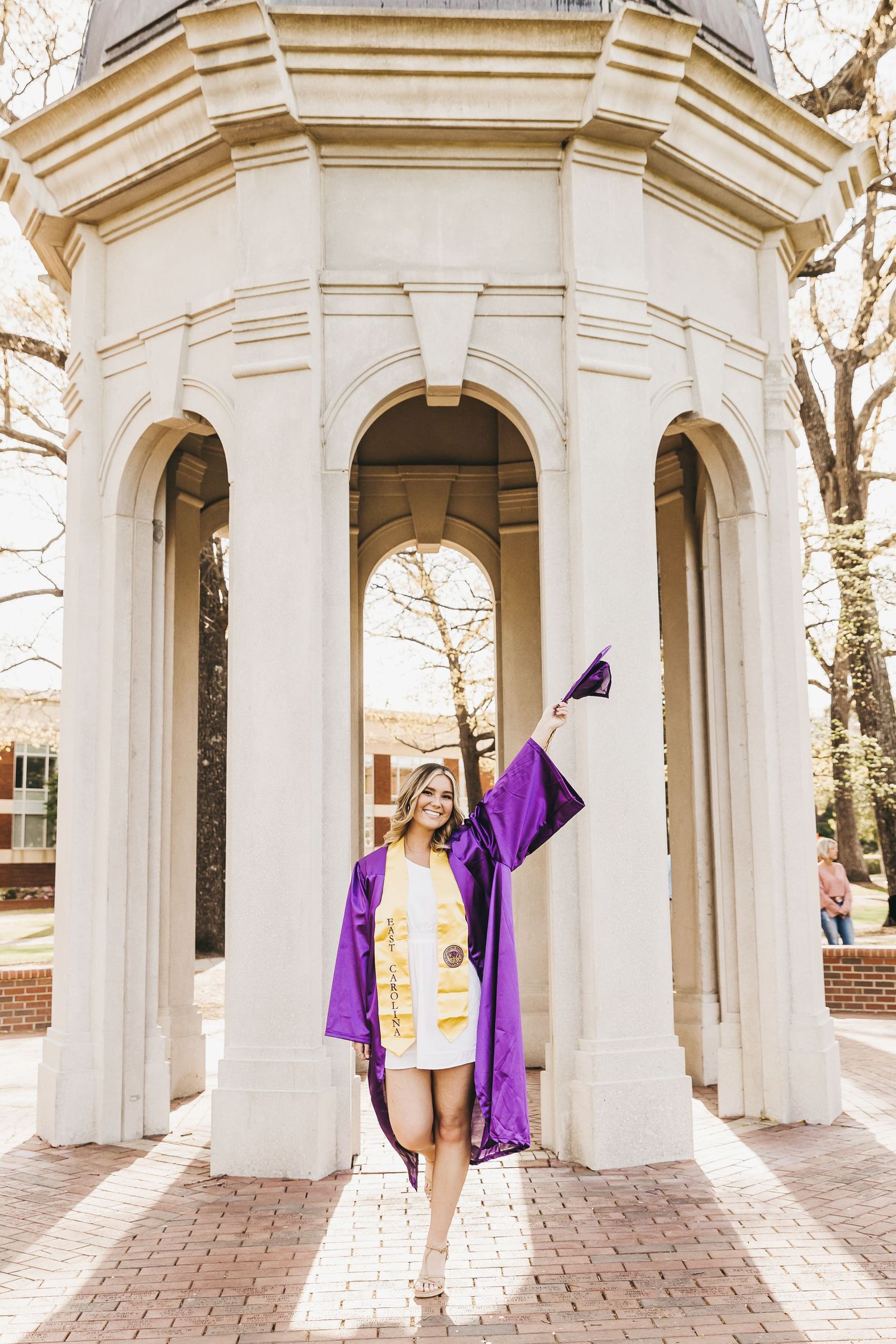 Graduate in purple gown and white dress celebrating under white architectural archway.