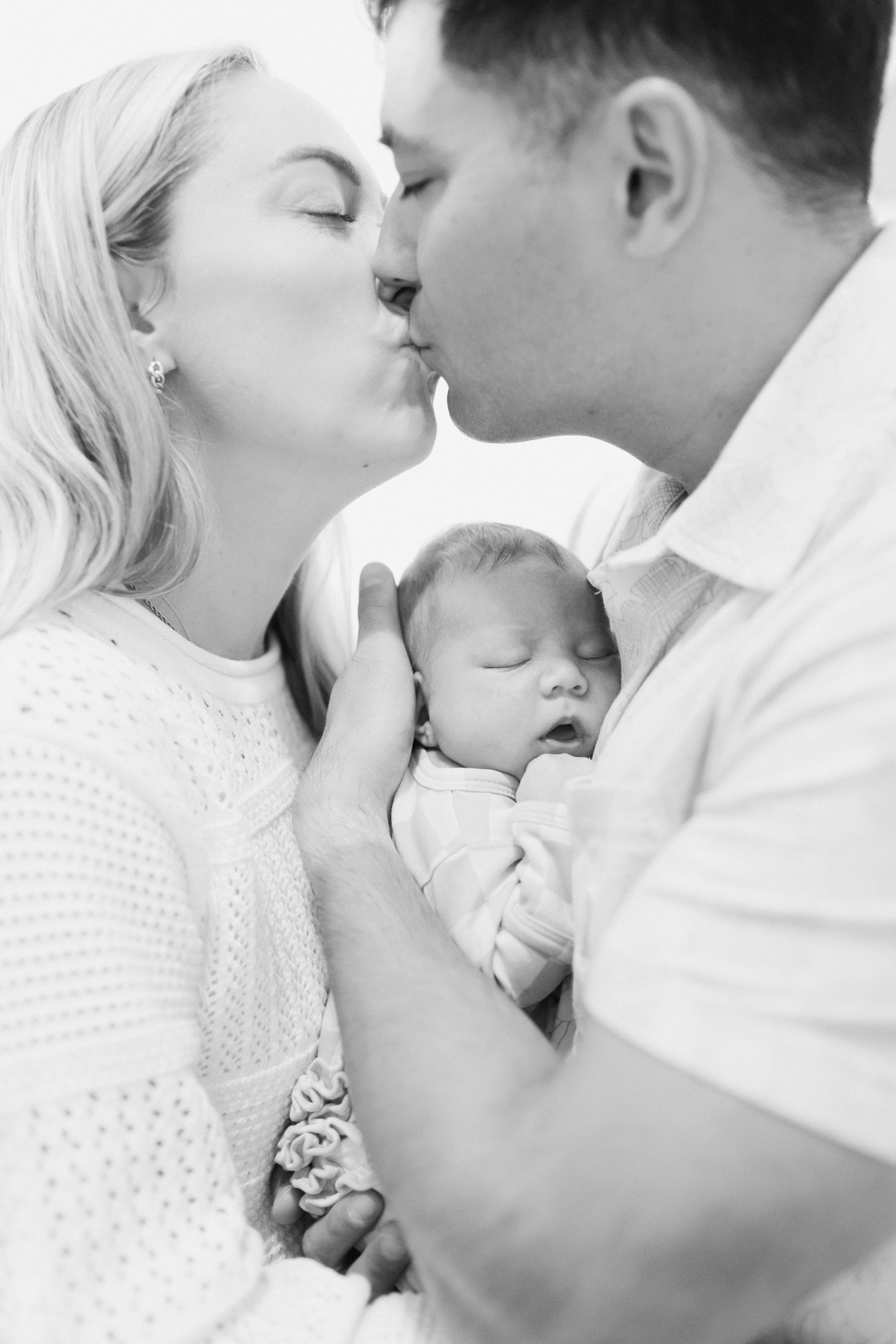 An intimate black and white portrait of a couple sharing a kiss while holding their newborn.