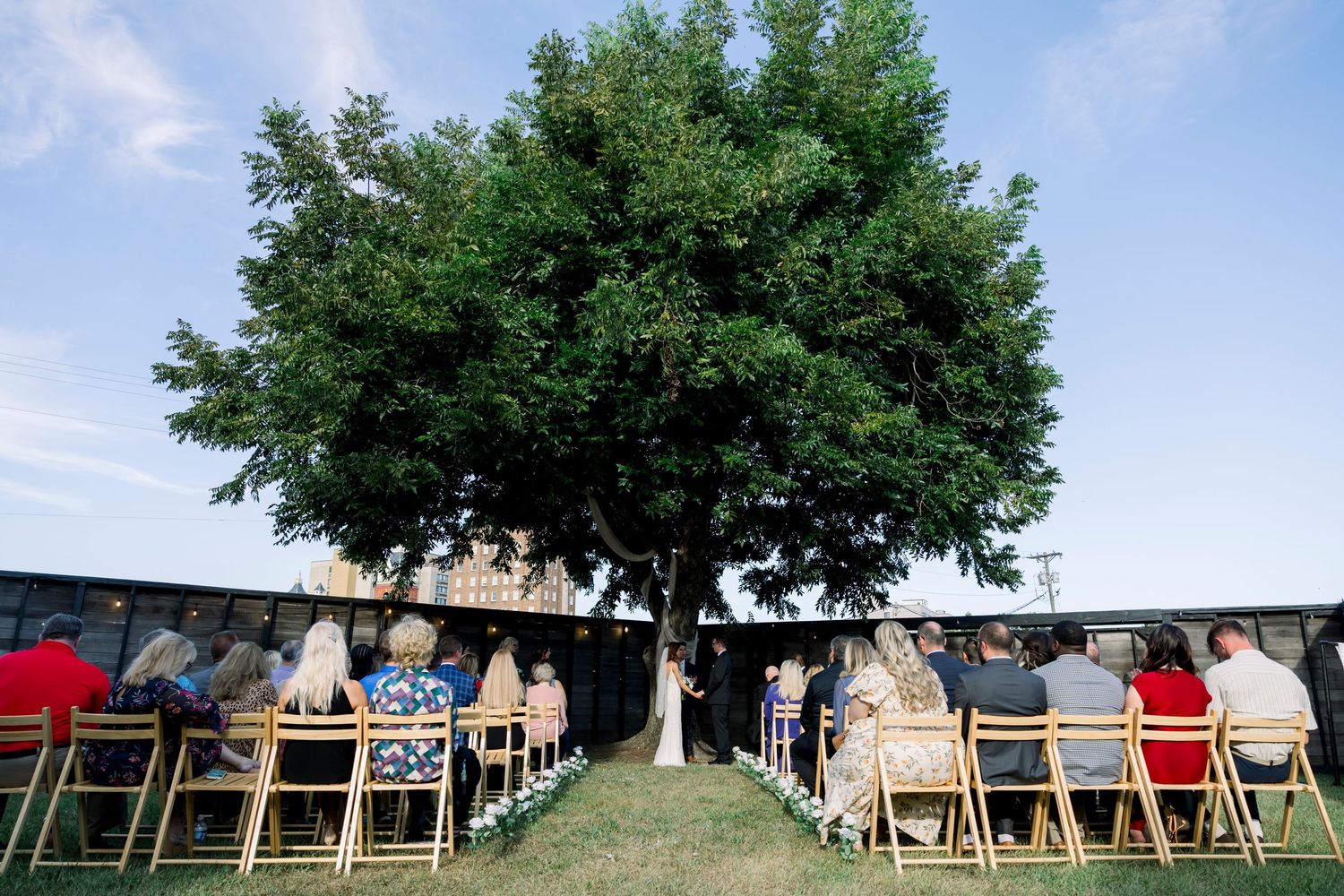Panoramic view of outdoor wedding ceremony setup with white chairs and trees in the background.