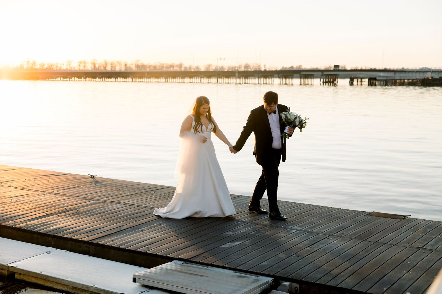 Couple in wedding attire walking along wooden dock during golden hour with water views.