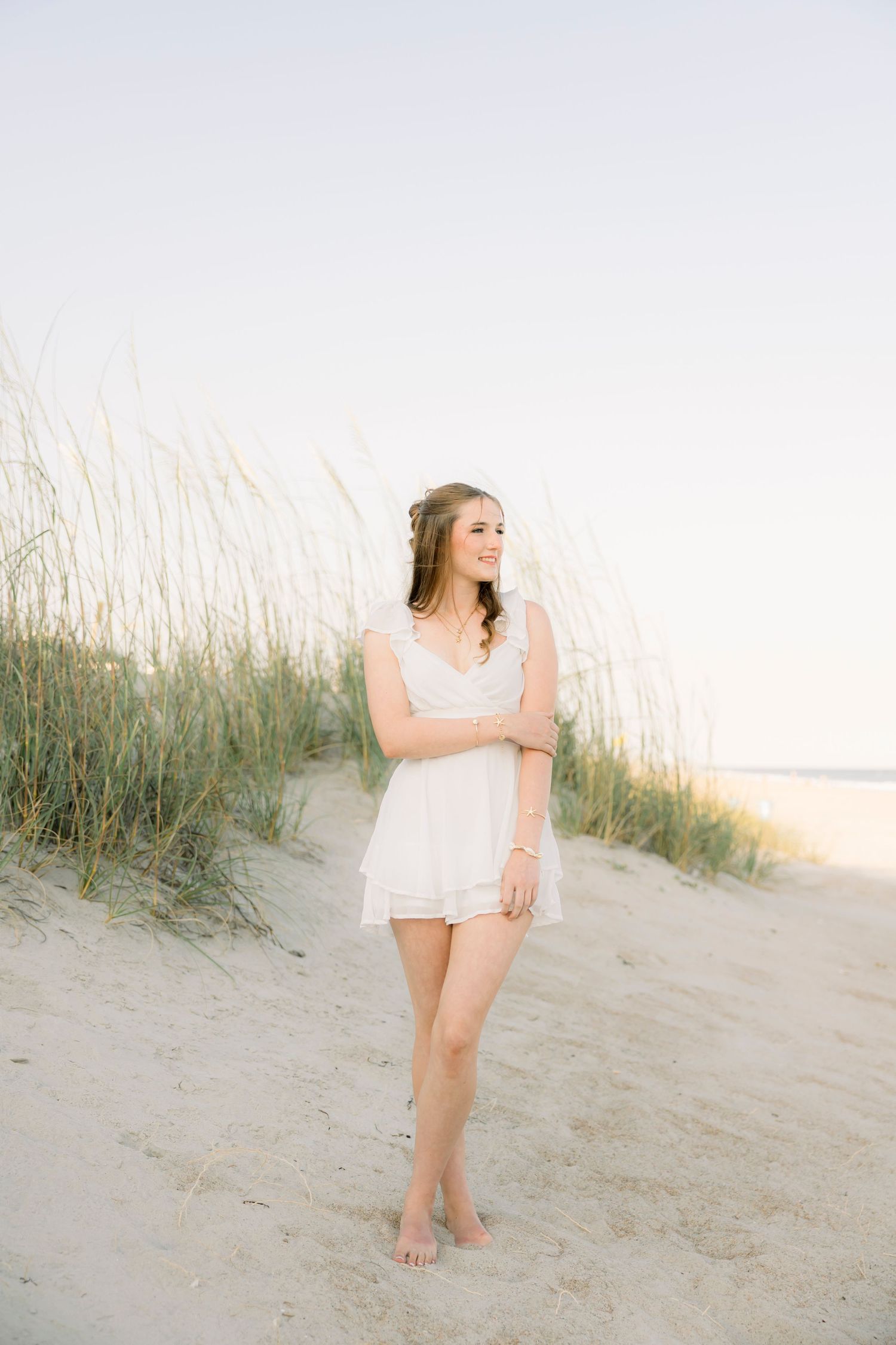Young woman in white dress standing barefoot on sandy beach dunes with tall grass, smiling in soft natural light.