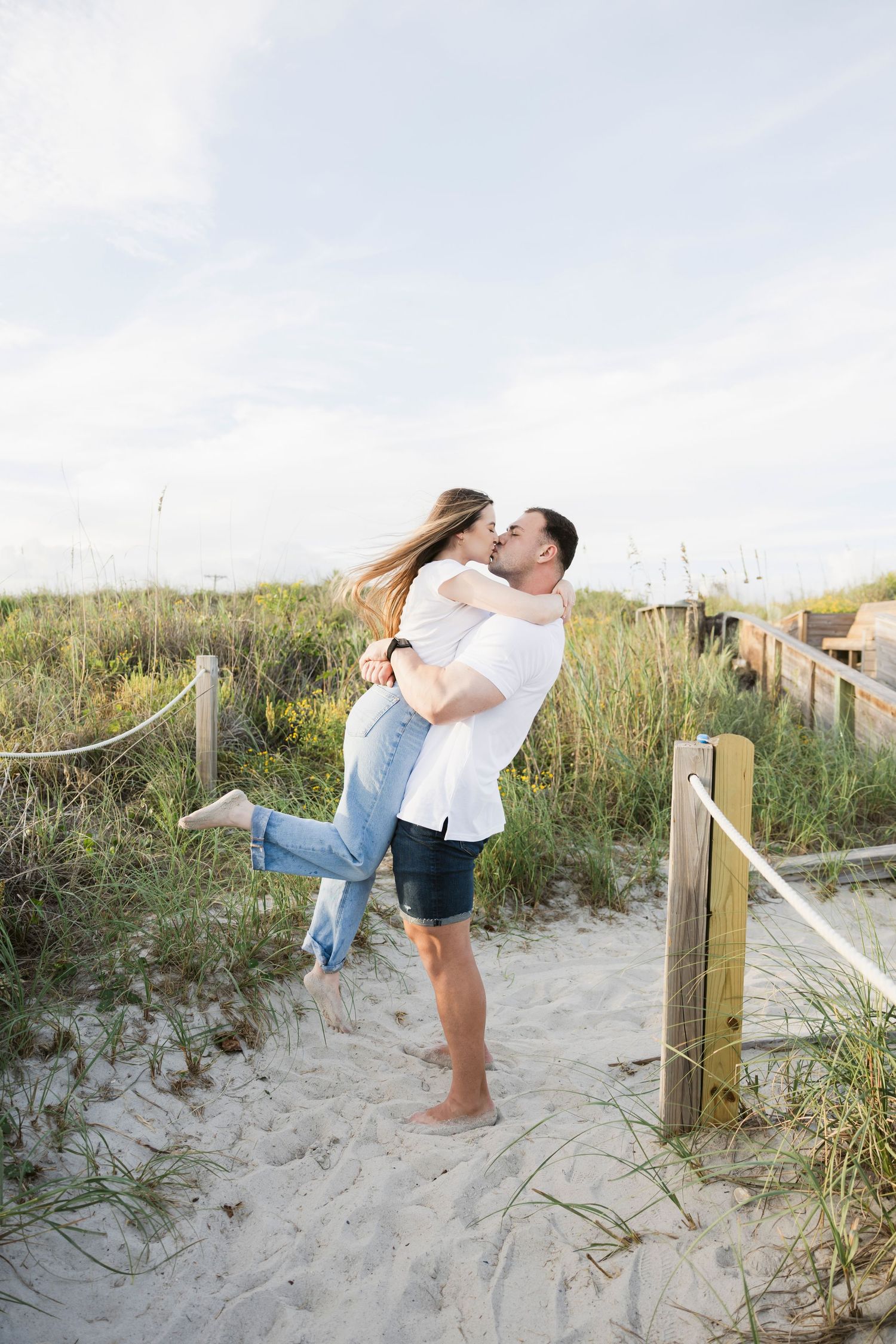 Playful couple embraces on sandy beach boardwalk during sunset photoshoot.