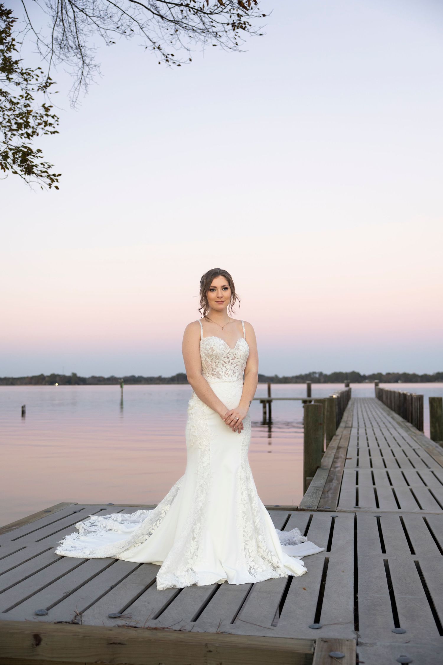 A white wedding dress with lace details photographed on a wooden dock at dusk.