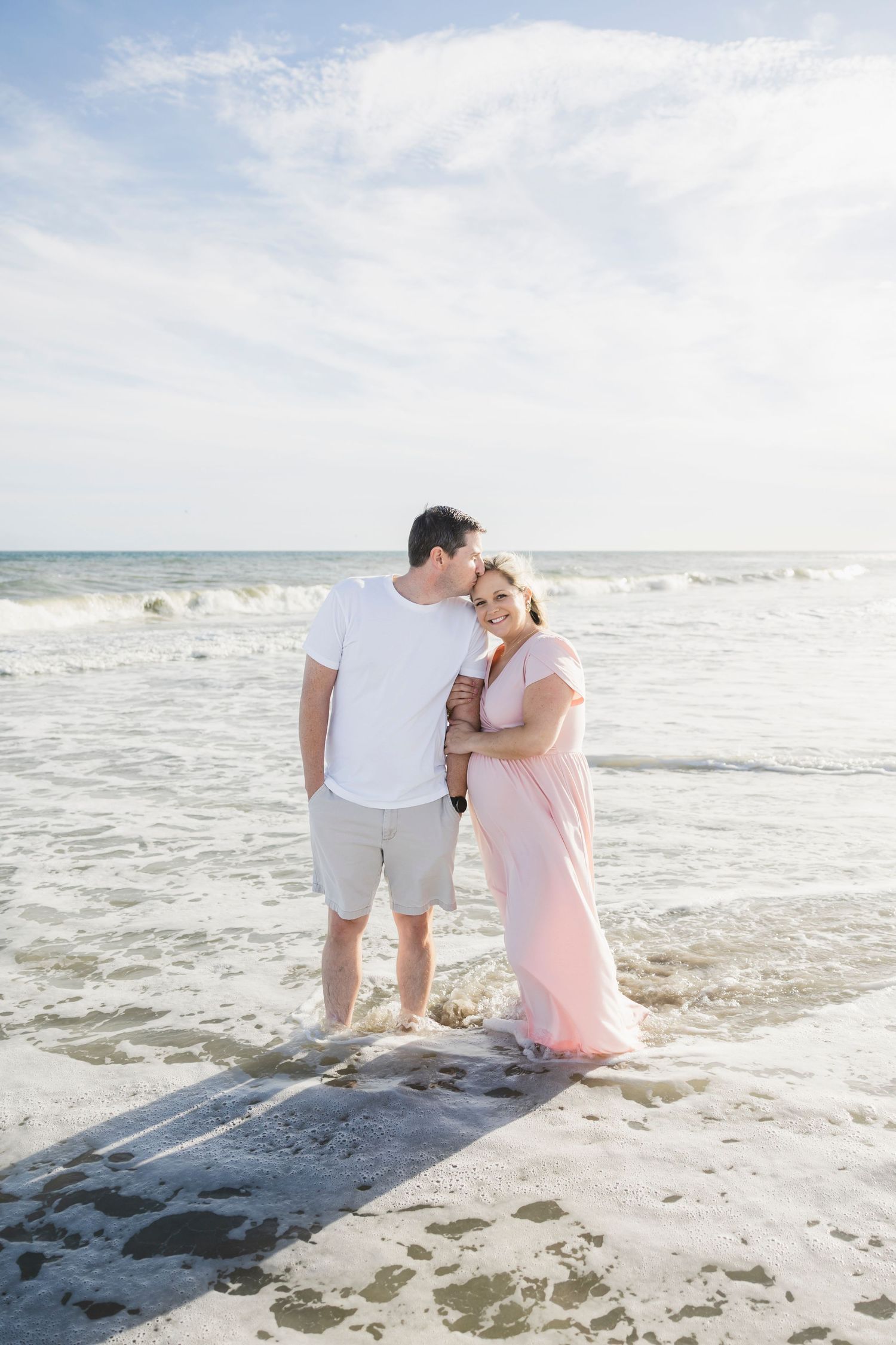 Couple shares tender moment during beach maternity shoot with ocean waves.