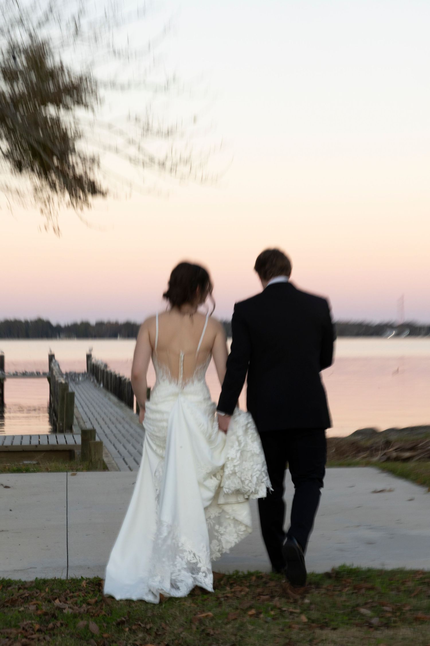 A couple in wedding attire walks along a waterfront boardwalk at sunset.