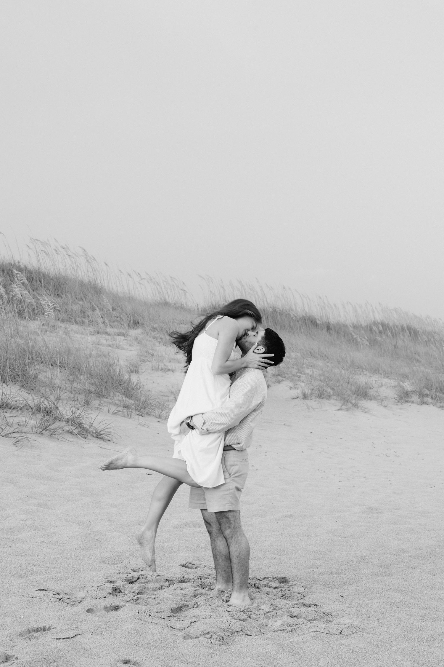 Black and white photo of a couple kissing on a sandy beach, man lifting woman as tall grass sways in the background.